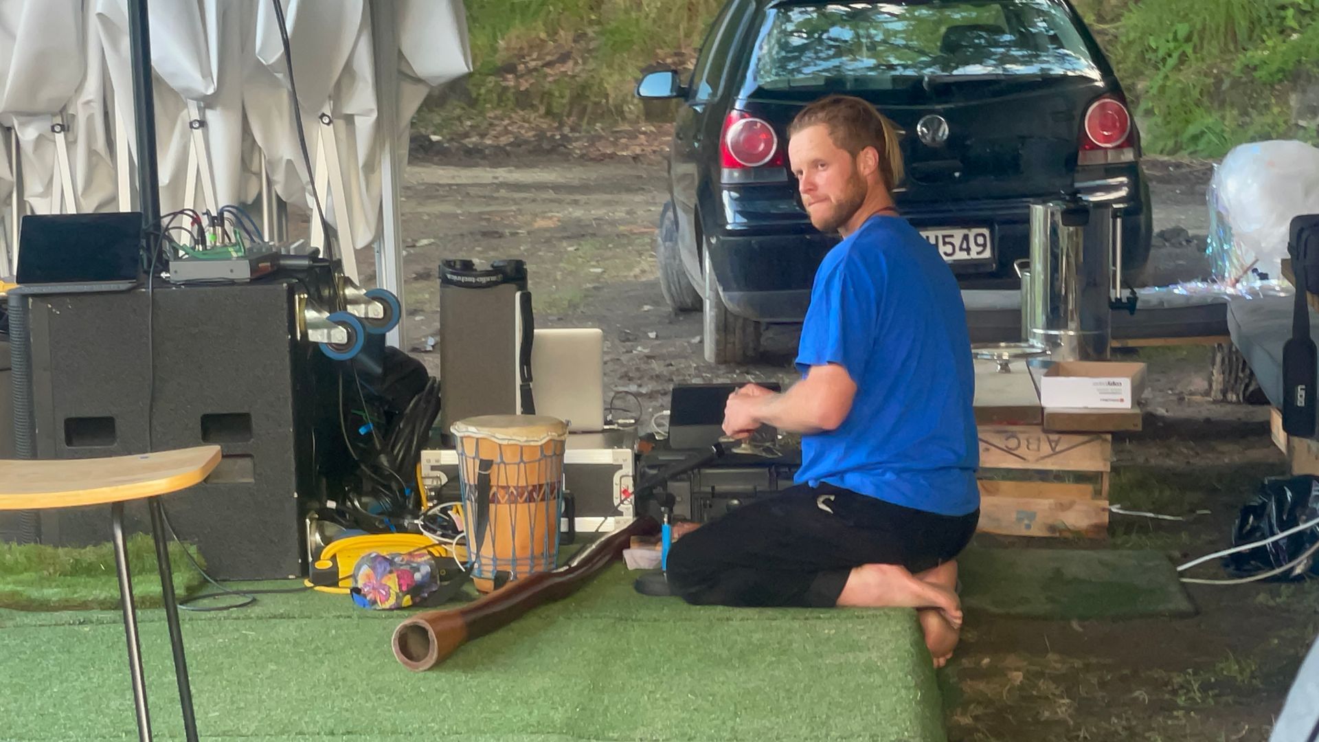 Man kneels, adjusting equipment near a drum and didgeridoo. A black car sits in background.
