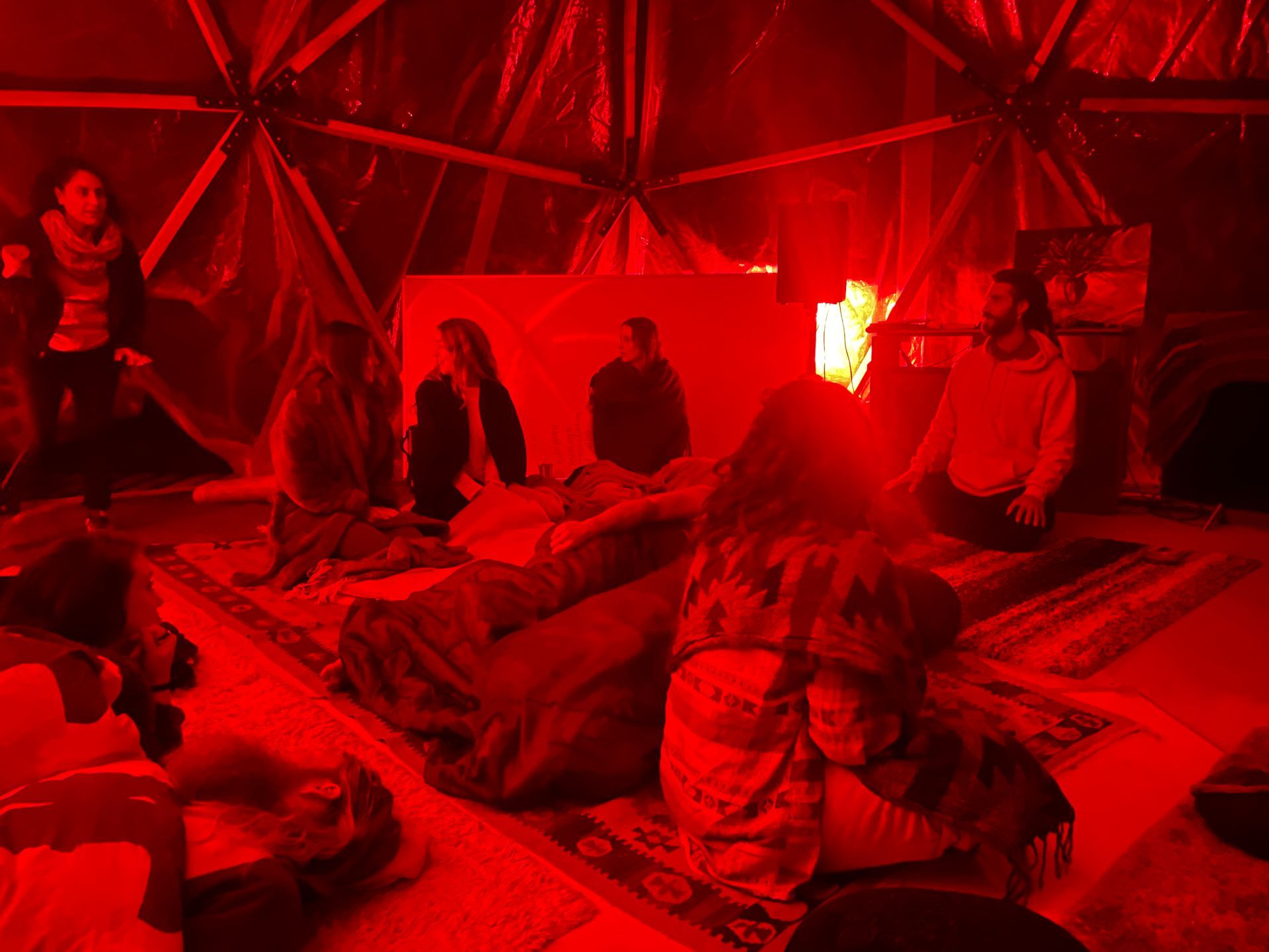People gather inside a red-lit dome. Some sit on a rug, others stand. A screen projects an image.