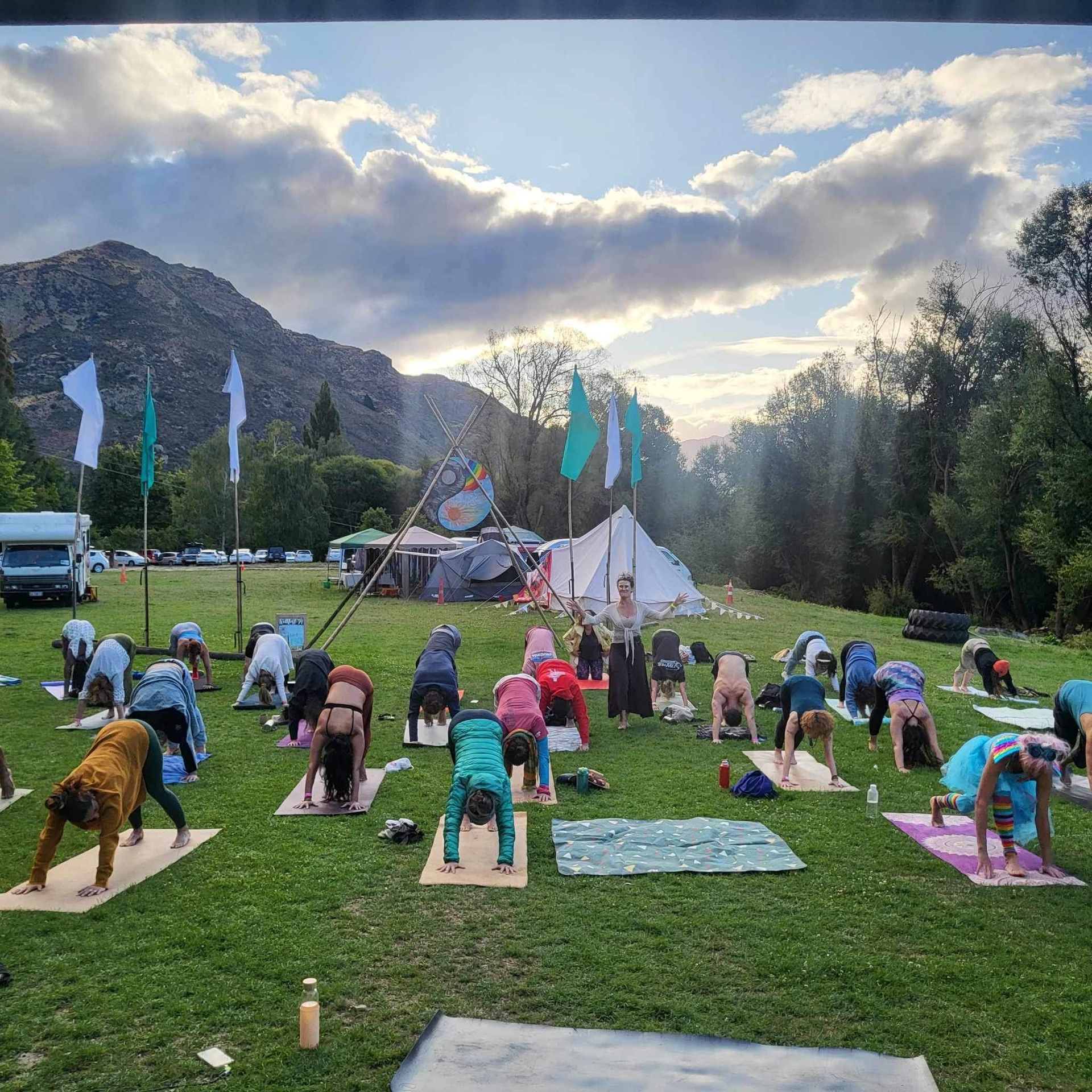 People doing yoga outdoors on a grassy field, mountains in the background, cloudy sky.