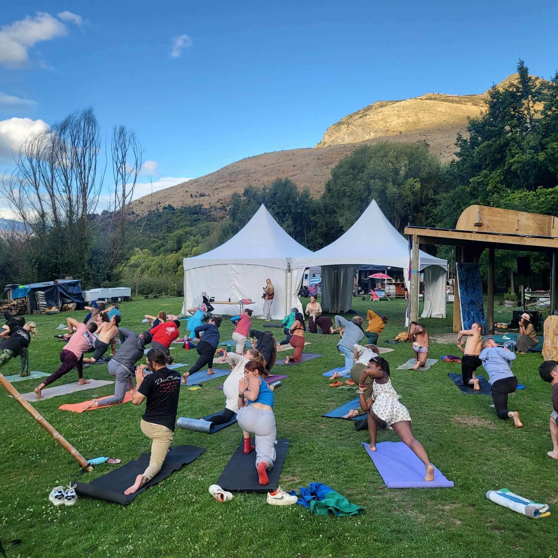 Large outdoor yoga class in a grassy park, mountain backdrop. People in various poses near two white tents.