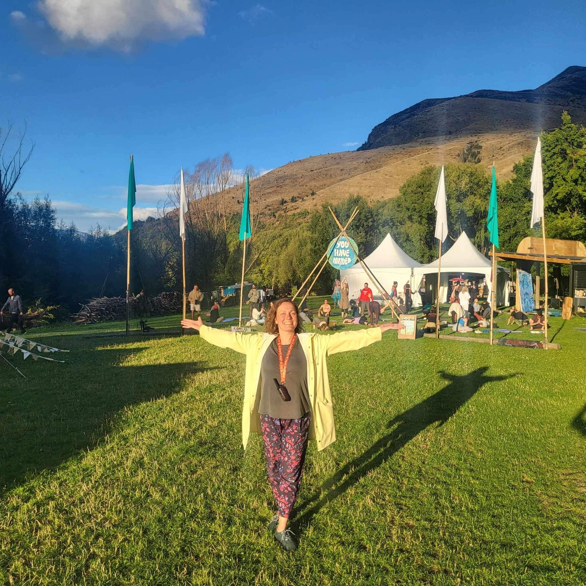 Woman with arms outstretched in a grassy field, tents and mountains in the background.
