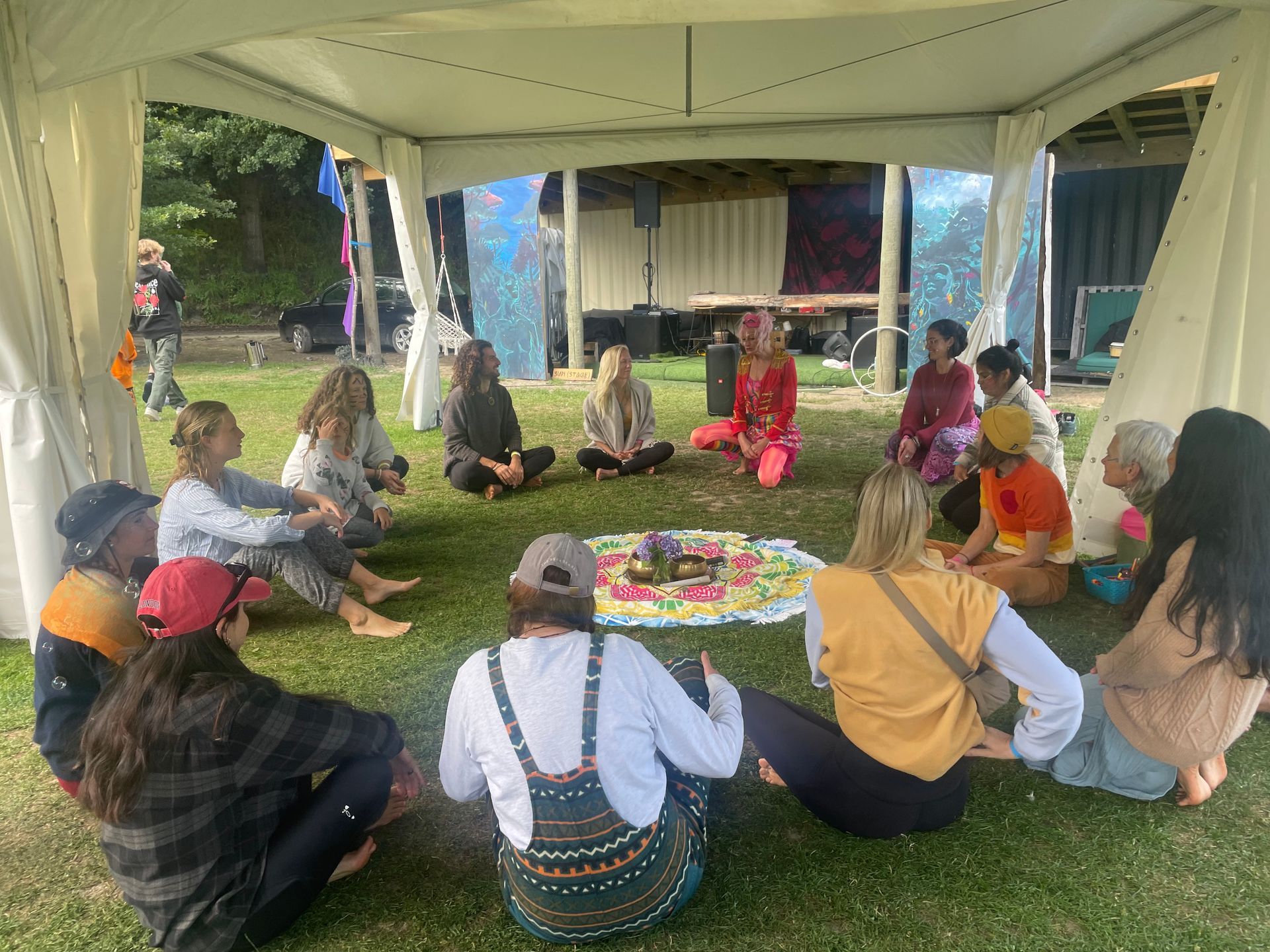 People seated in a circle on grass under a tent. A colorful arrangement is in the center.