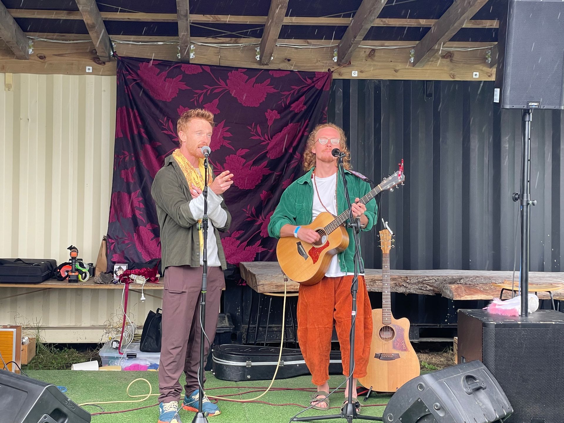 Two musicians perform on a stage outdoors. One sings and plays guitar, the other sings and claps. Purple backdrop.