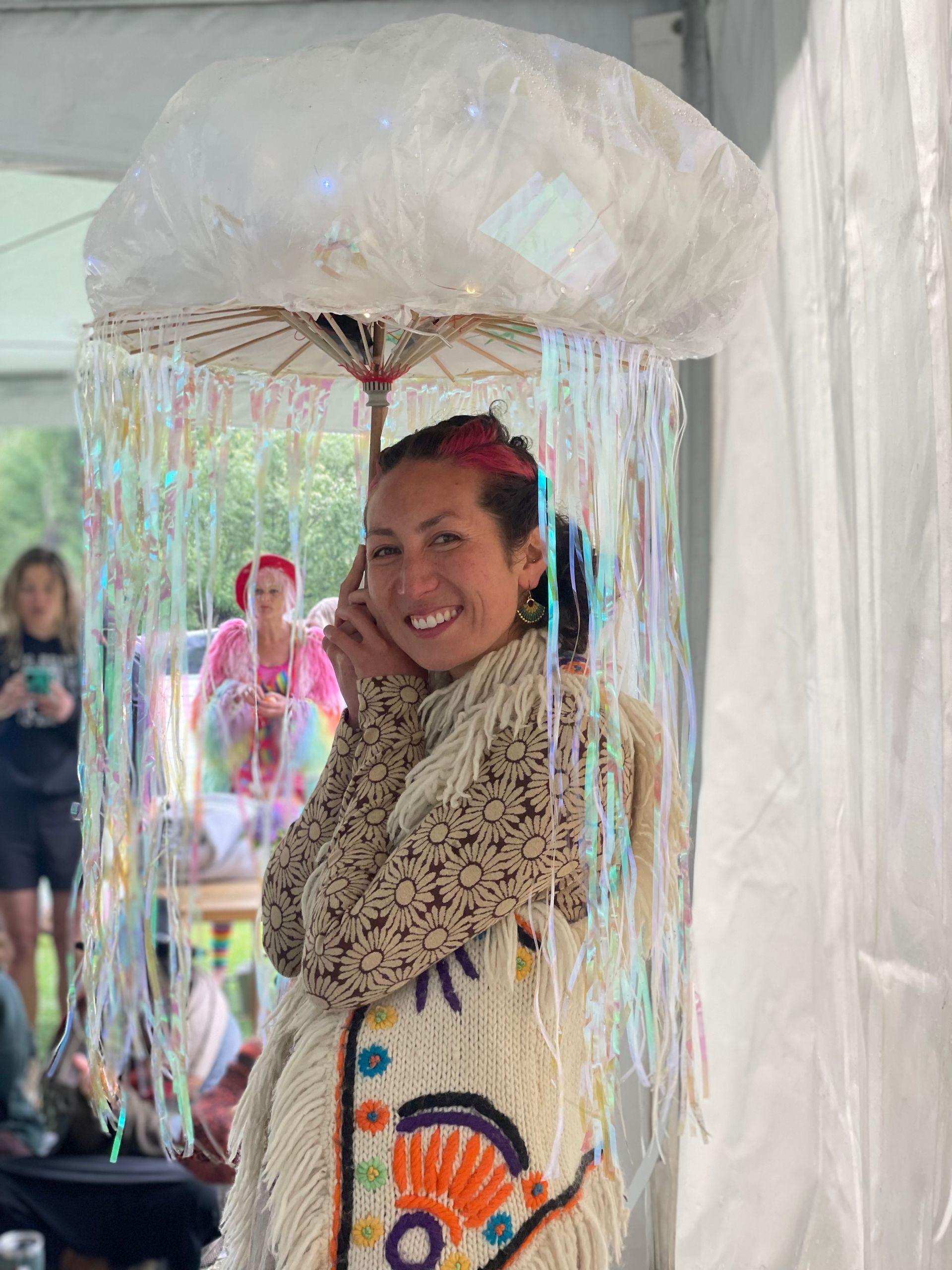 Woman wearing cloud-shaped hat with shimmering streamers and decorative vest smiles. Outdoors setting.
