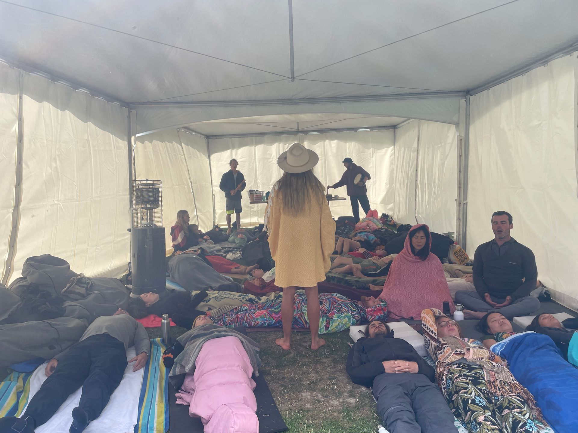 People resting on mats inside a tent; a person stands wearing a hat. Two others stand in the background.