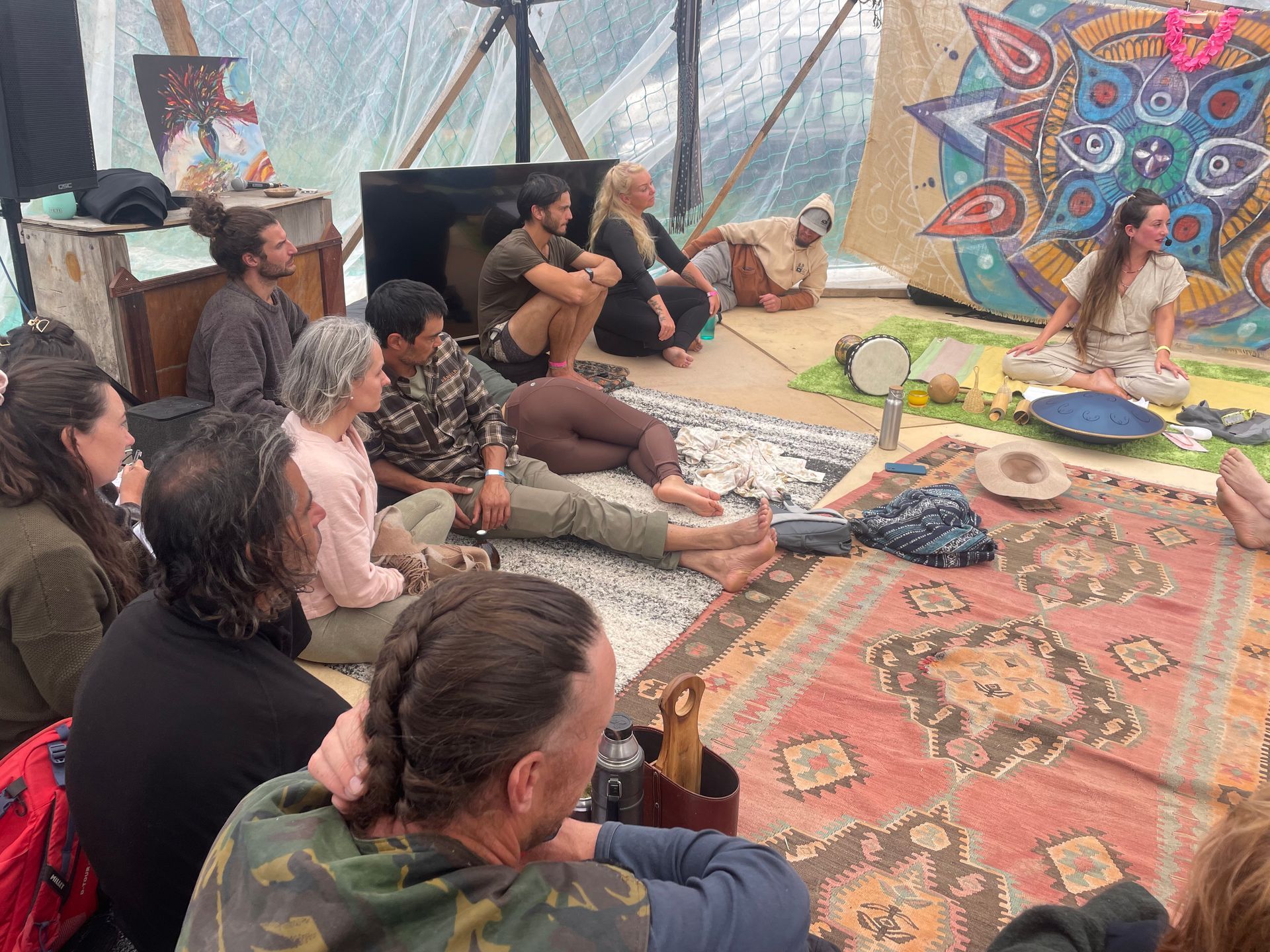 People seated on rugs and a platform, watching a woman speaking and playing instruments inside a tent.