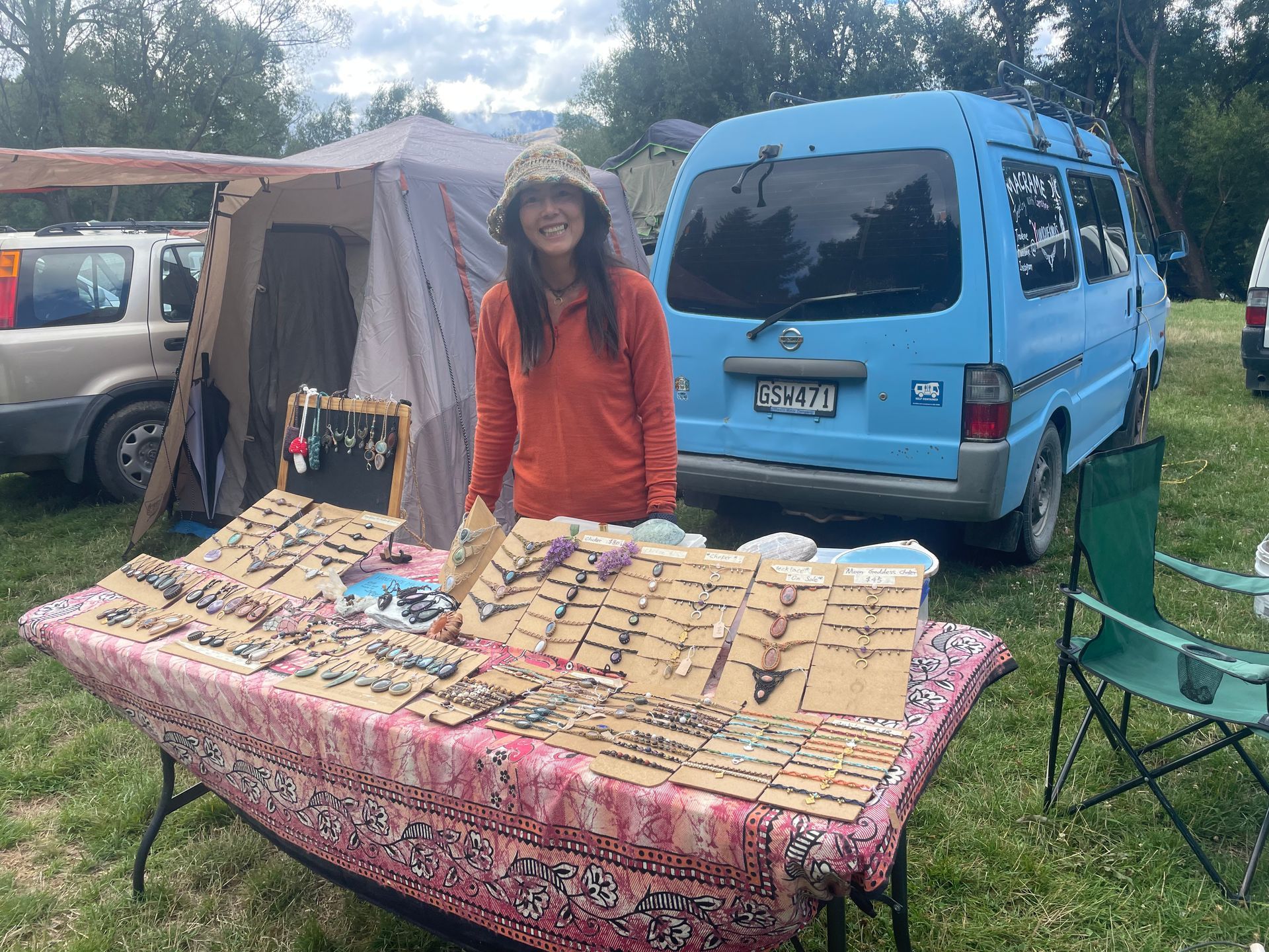 Woman at a craft market with jewelry on display in front of a blue van.