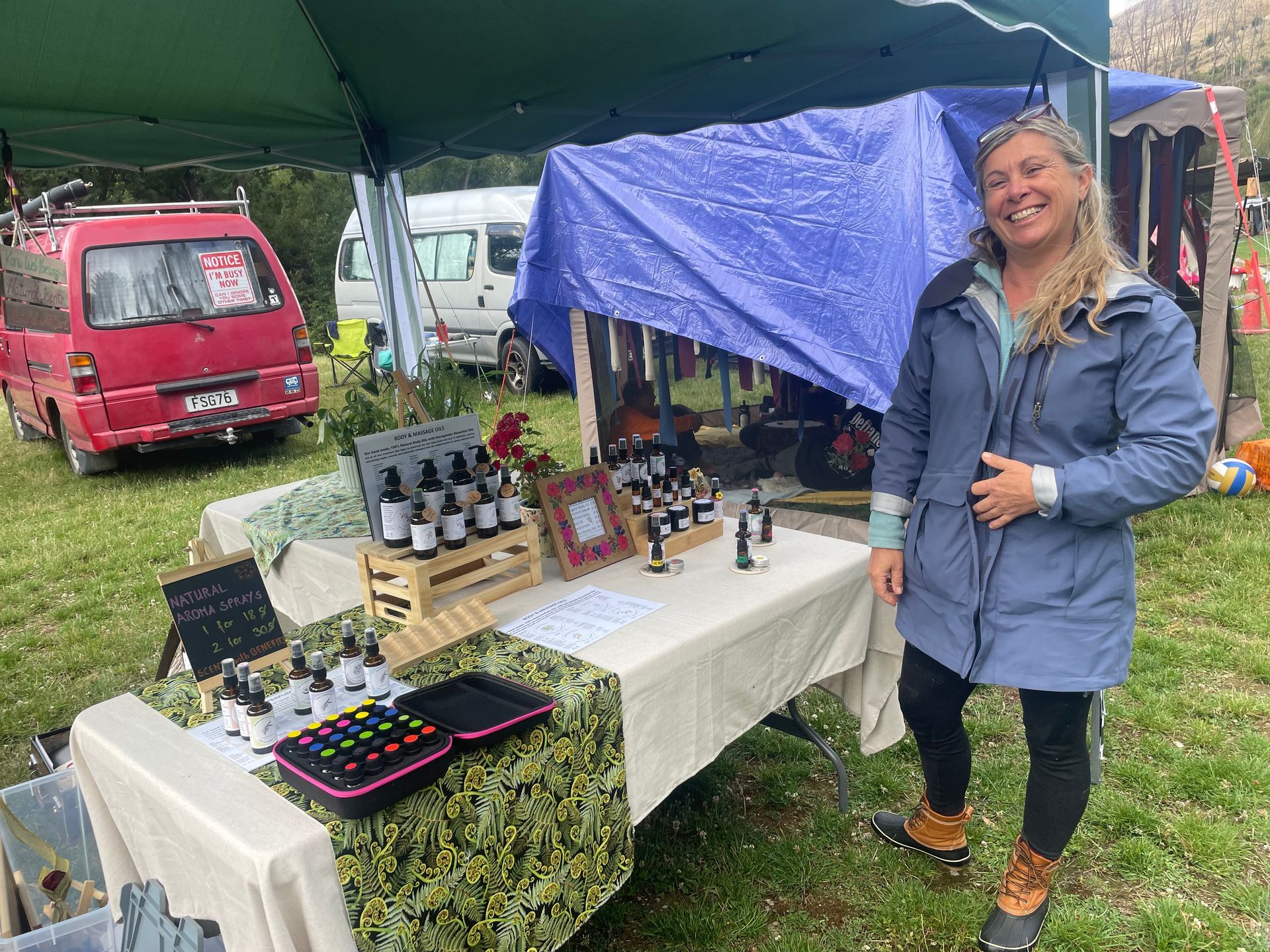 Woman smiles at a table of products at an outdoor market, beside small vans and a tent.