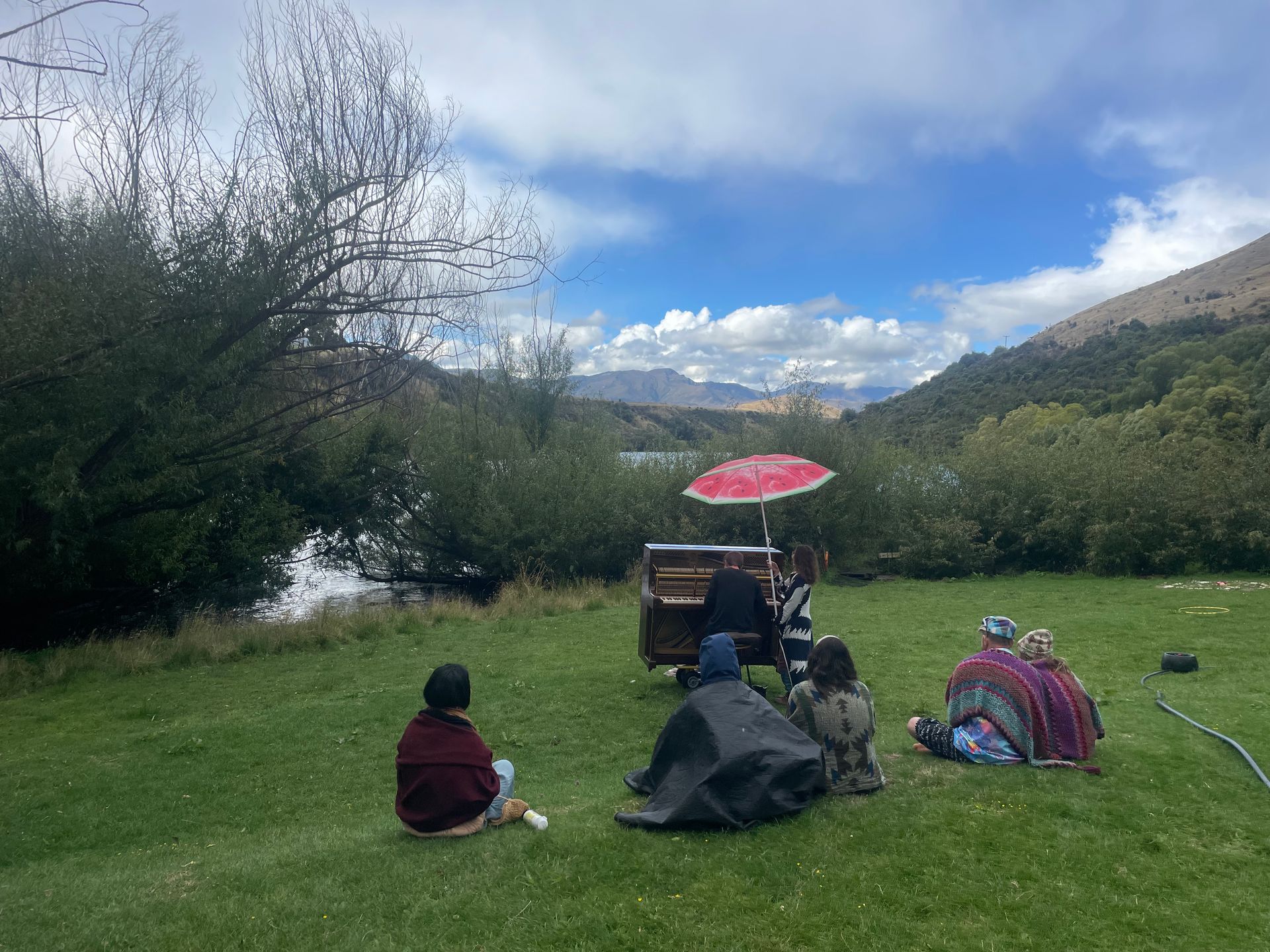 People gathered near a food cart under a red umbrella on a grassy field, near water, with mountains in the distance.