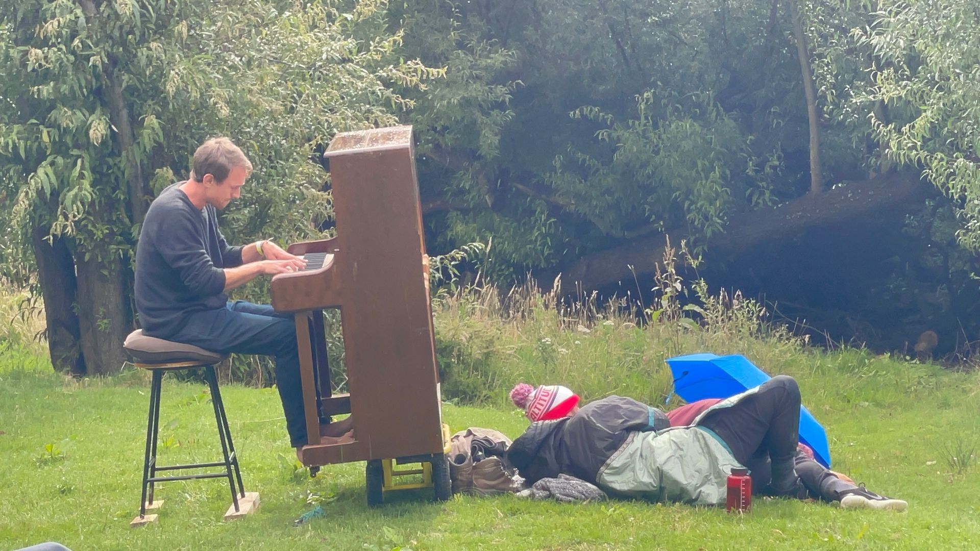 Man playing piano outdoors, seated on a stool. Someone reclines on the grass nearby, surrounded by belongings.