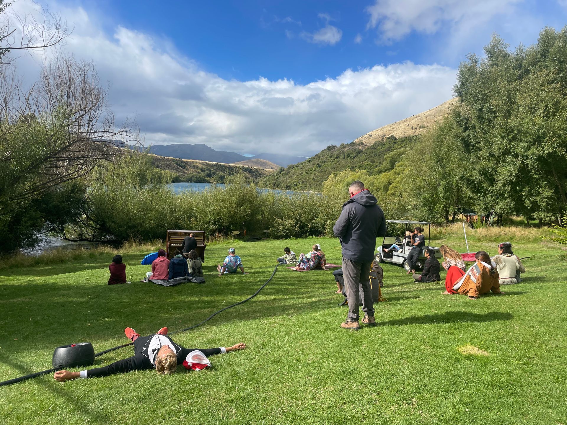 People gathered outdoors on a grassy lawn with a lake and mountains in the background. A man stands, and someone lies on the grass.