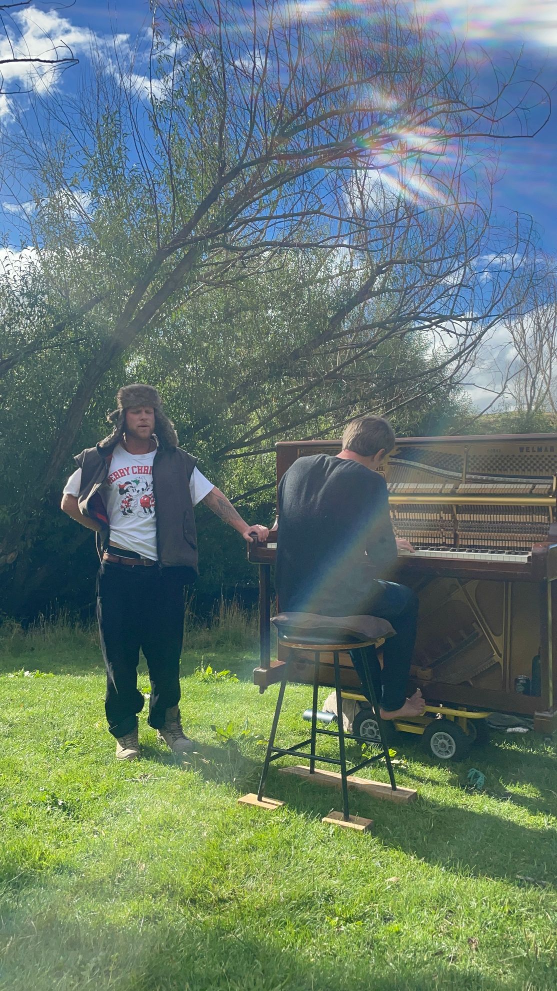 Two people playing a piano outdoors. One stands, wearing a vest and hat. The other sits, playing.