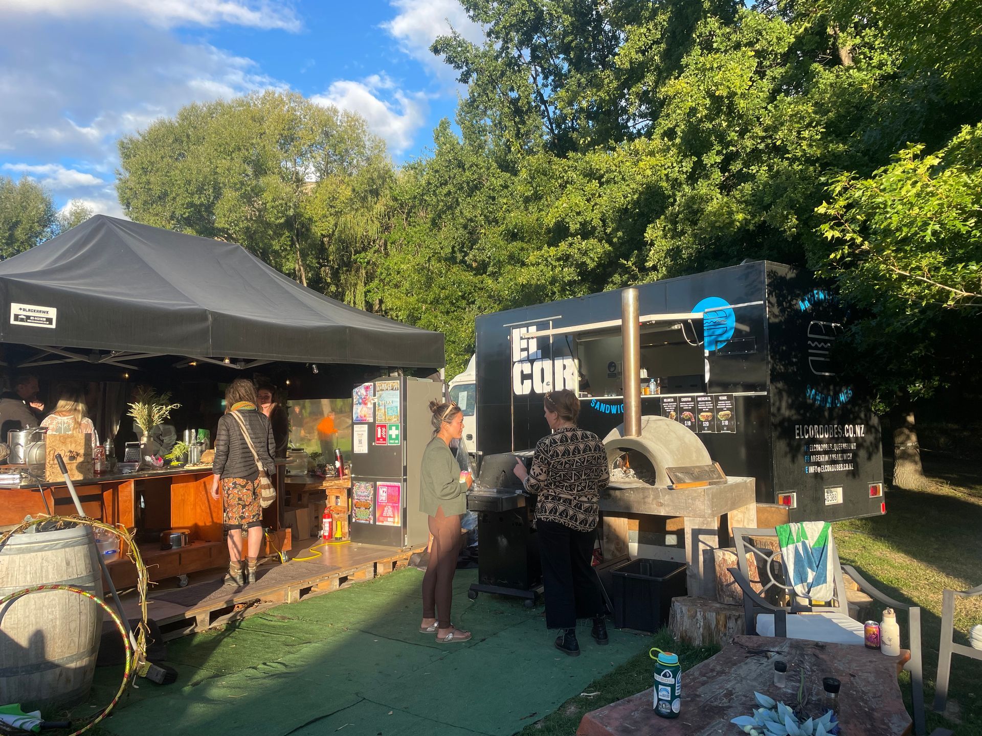 Food vendors at outdoor event: black tent and trailer with wood-fired pizza oven; people order food in park setting.