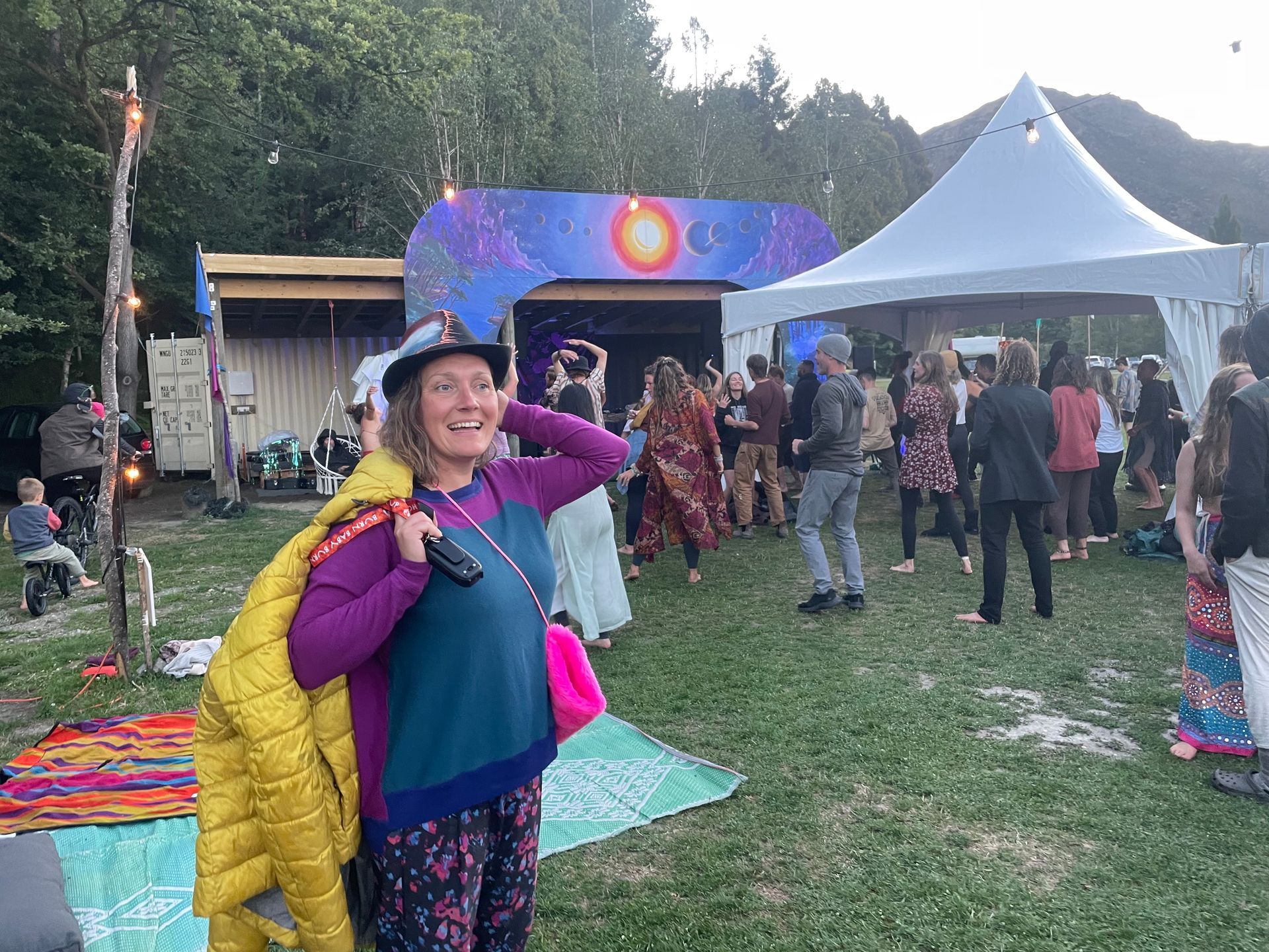 Woman smiling, holding jacket, at outdoor event with stage and dancing crowd.