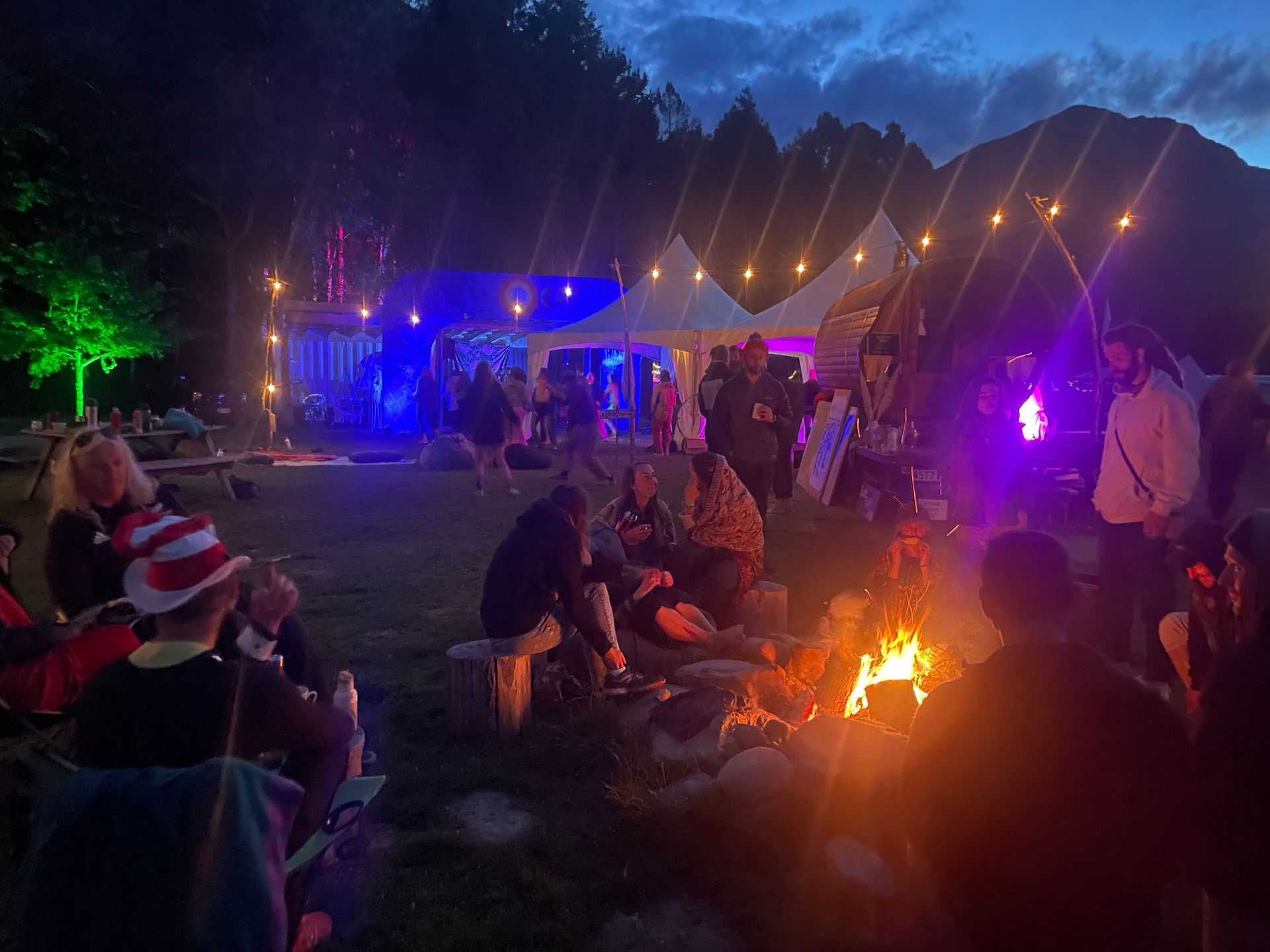 Nighttime outdoor gathering with bonfire, tents, and people socializing.