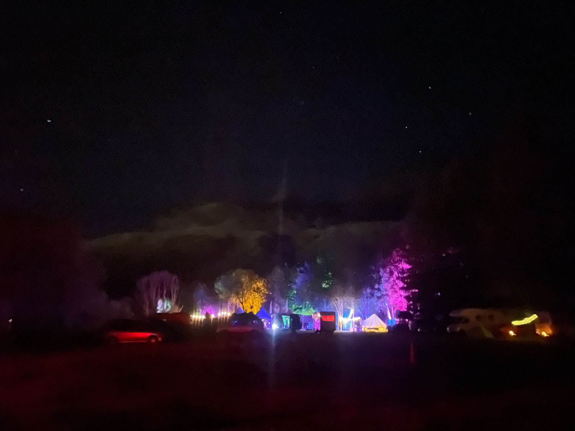 Night campsite with colorful lights illuminating trees and tents under a starry sky.