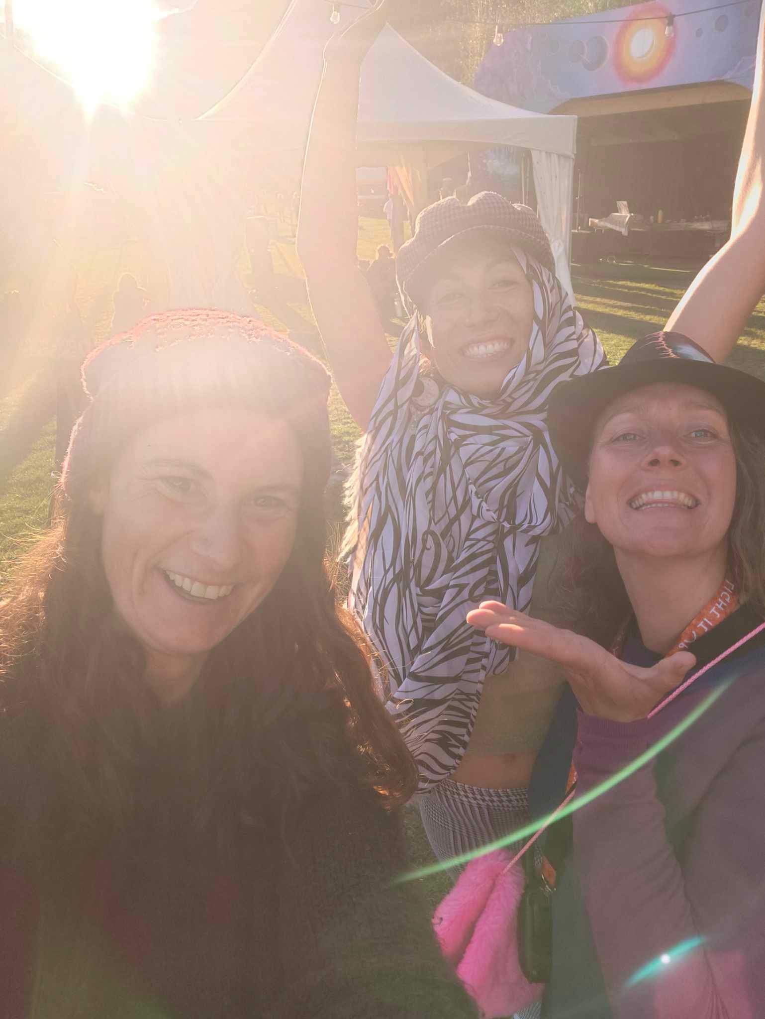 Three smiling people outdoors, arms raised, sunlit with a tent in background.