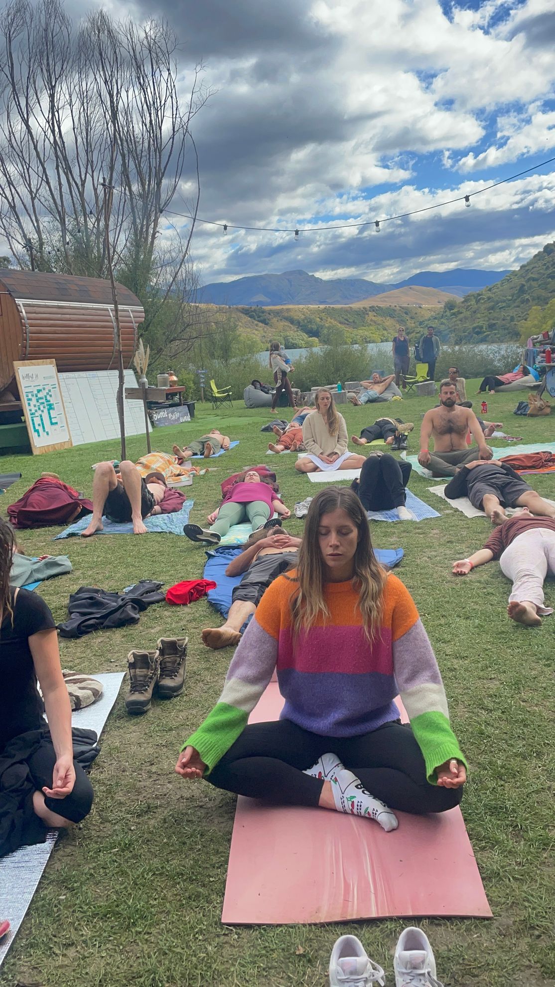 Group of people doing yoga on grass. Woman in sweater meditates; others recline. Outdoors with mountains.