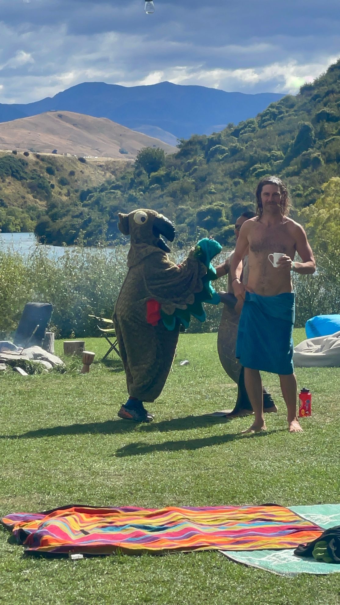 Person wrapped in a towel, dances with someone in a bird costume on a grassy lawn by a lake.