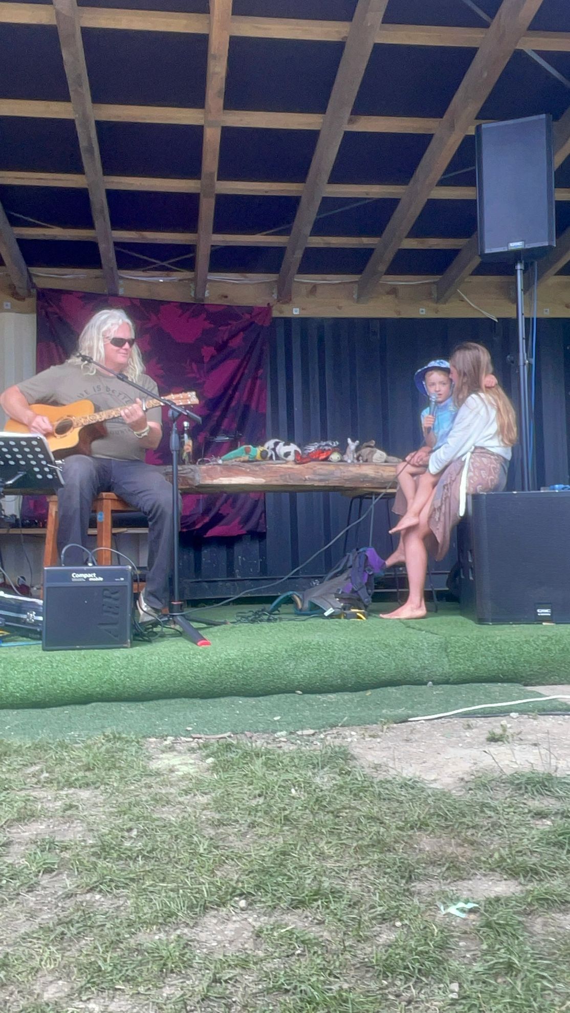 Man playing guitar, seated, performing outdoors; woman and child seated nearby.