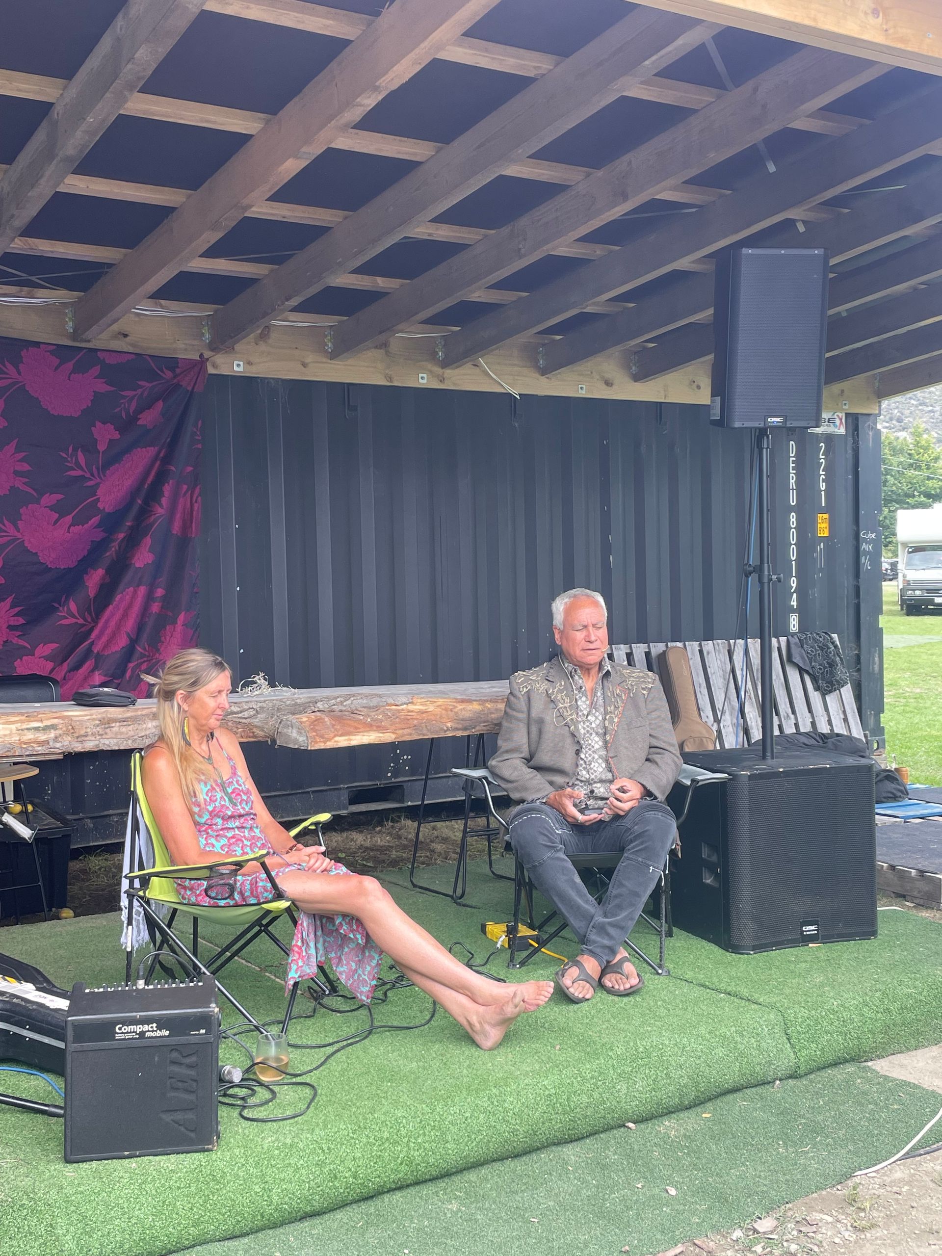Woman and man seated on a grassy stage under a wooden canopy, speaking.