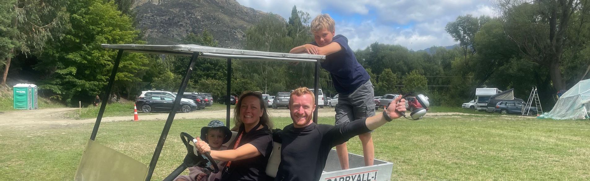 A family of four poses in a golf cart in a grassy field. A young boy leans over the top. Mountain backdrop.
