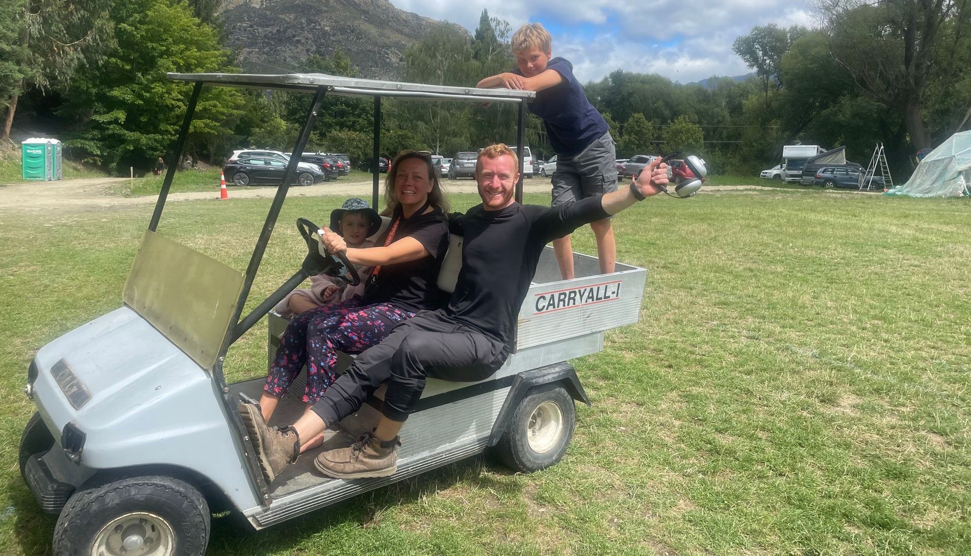 Family on a gray golf cart in a grassy area, a mountain in the background. Two adults and two children smile.