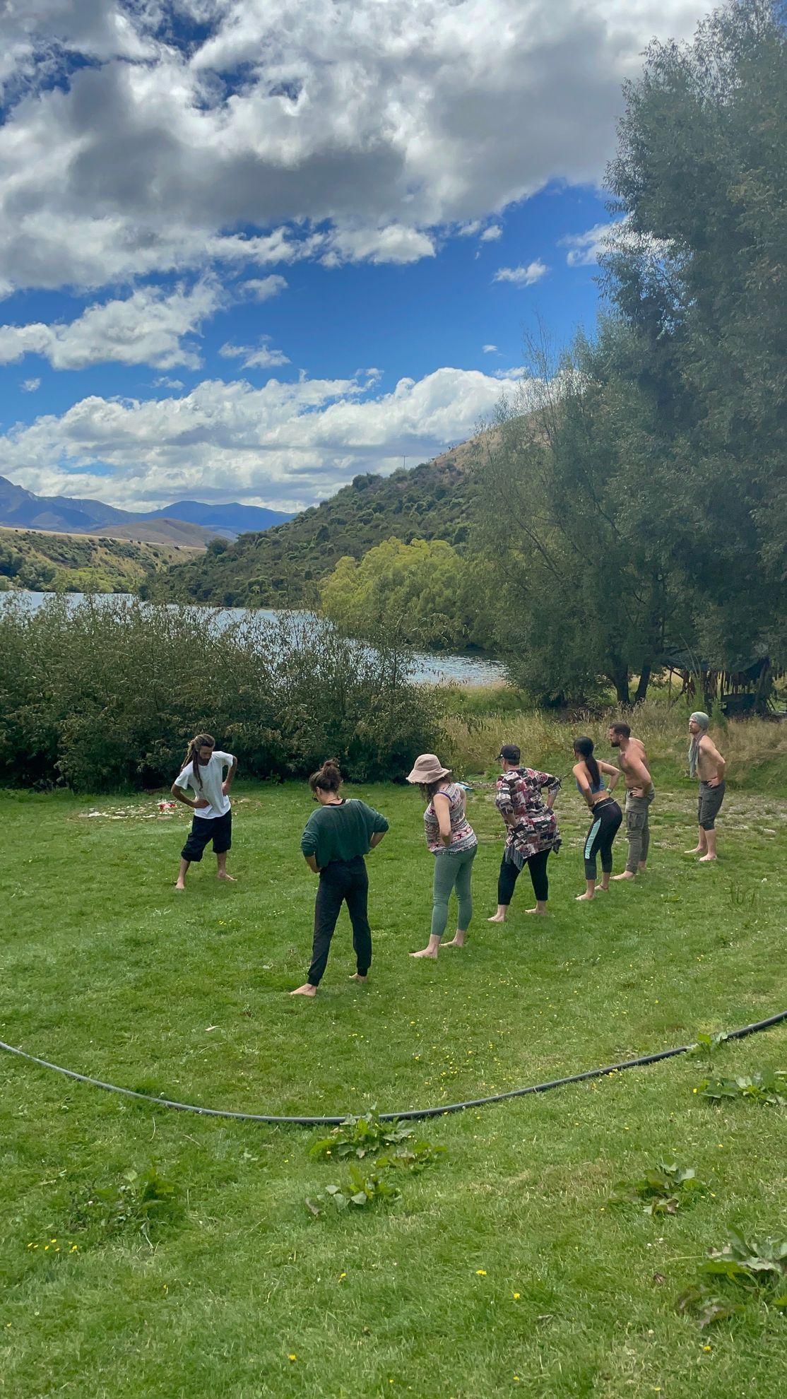 People stand in a grassy circle near a lake and trees under a blue sky.