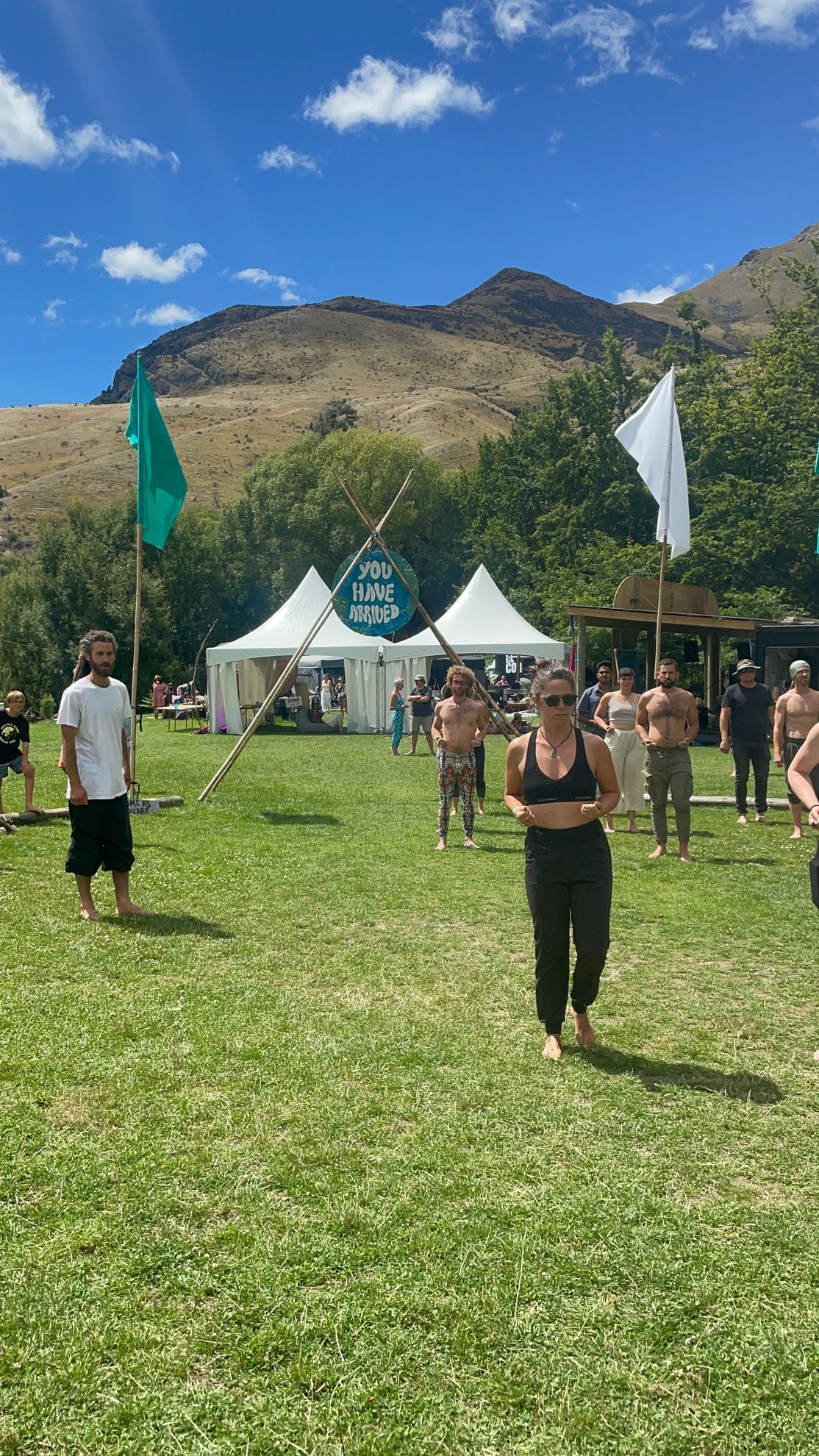 People doing outdoor yoga, tents, flags, mountains, blue sky, and green grass.