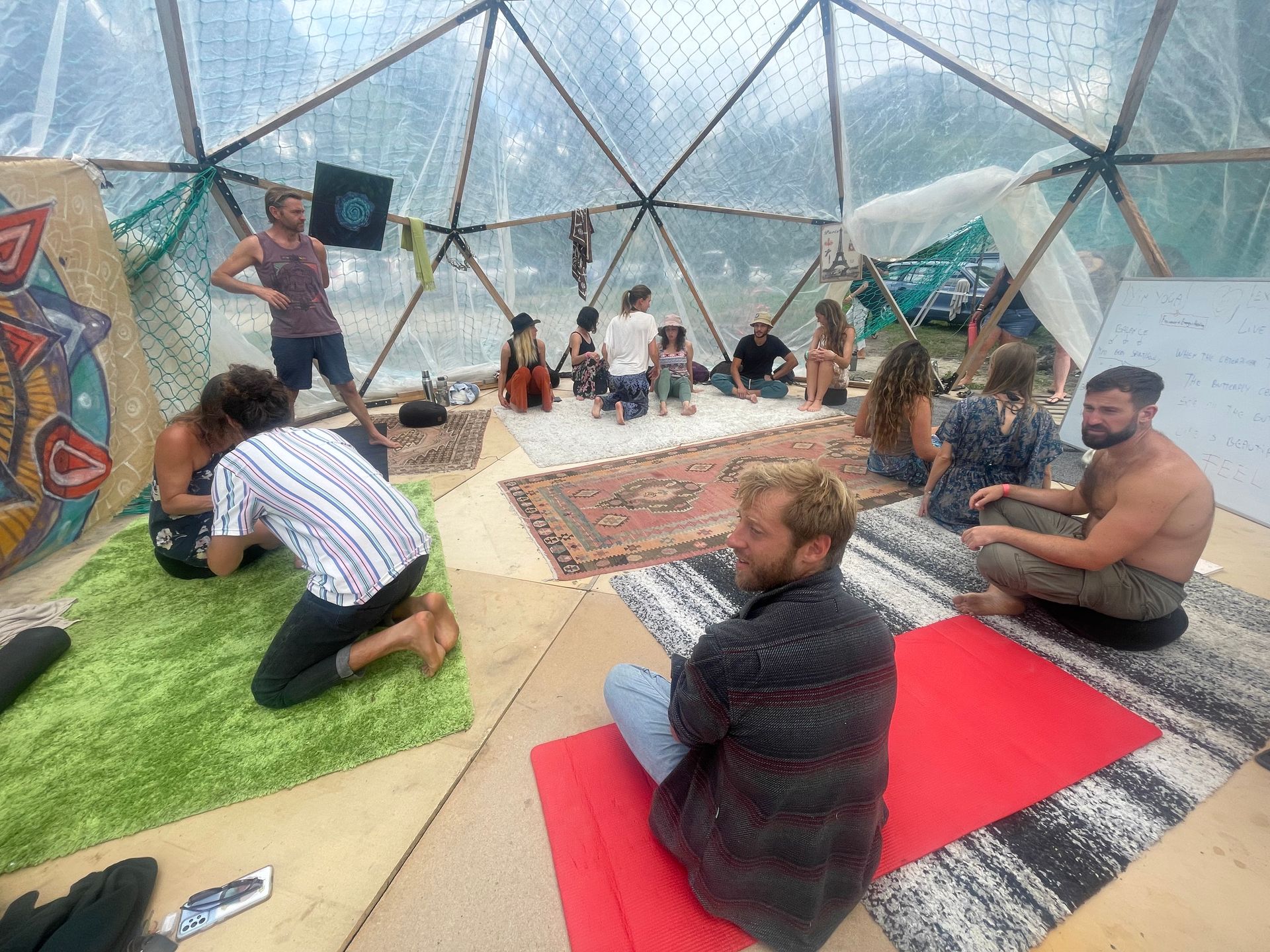 People in a clear dome, some seated on rugs or mats, others standing.
