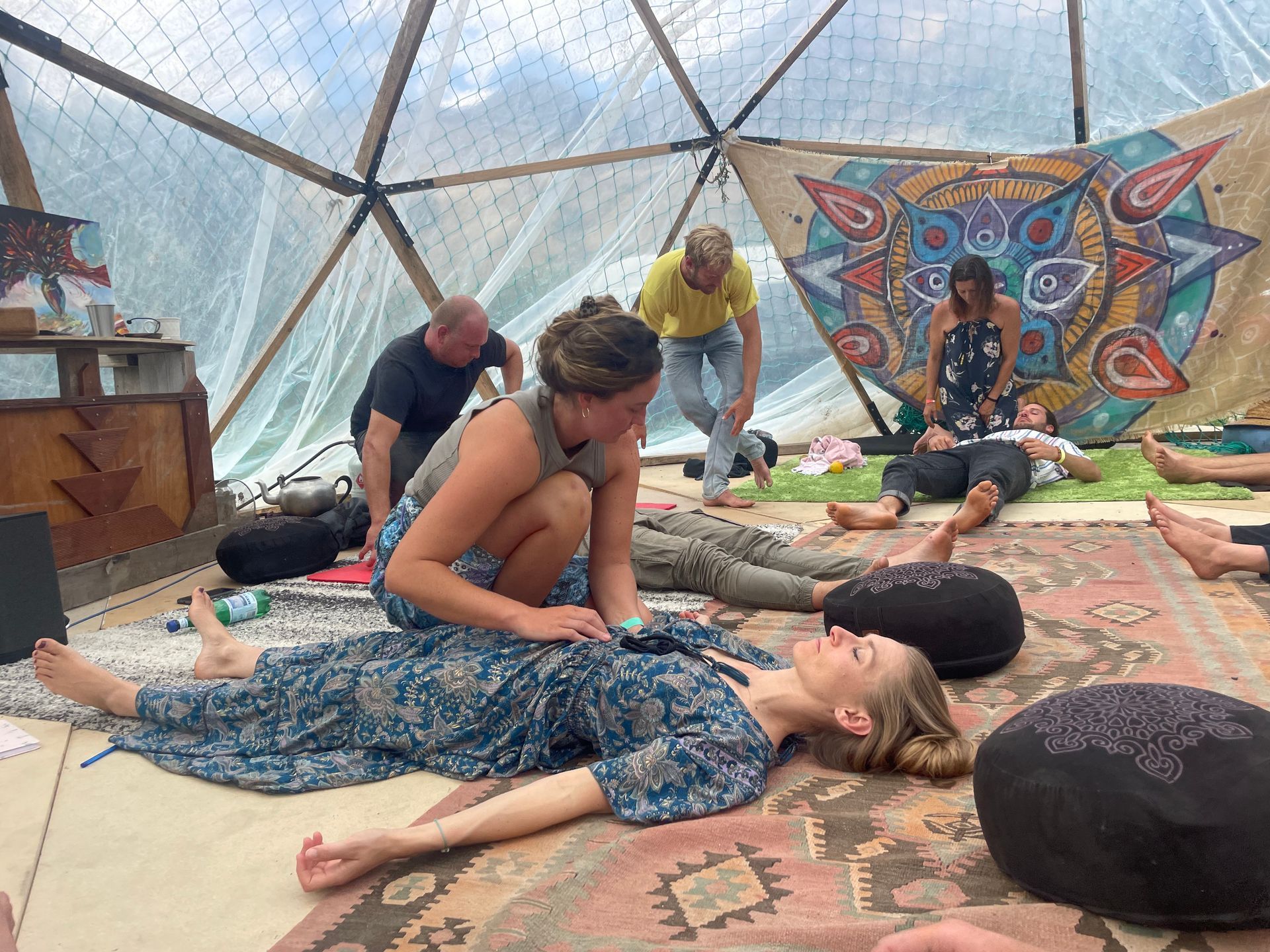 Group therapy session inside a dome. Participants lying down, some receiving hands-on treatment. Colorful rug and artwork present.