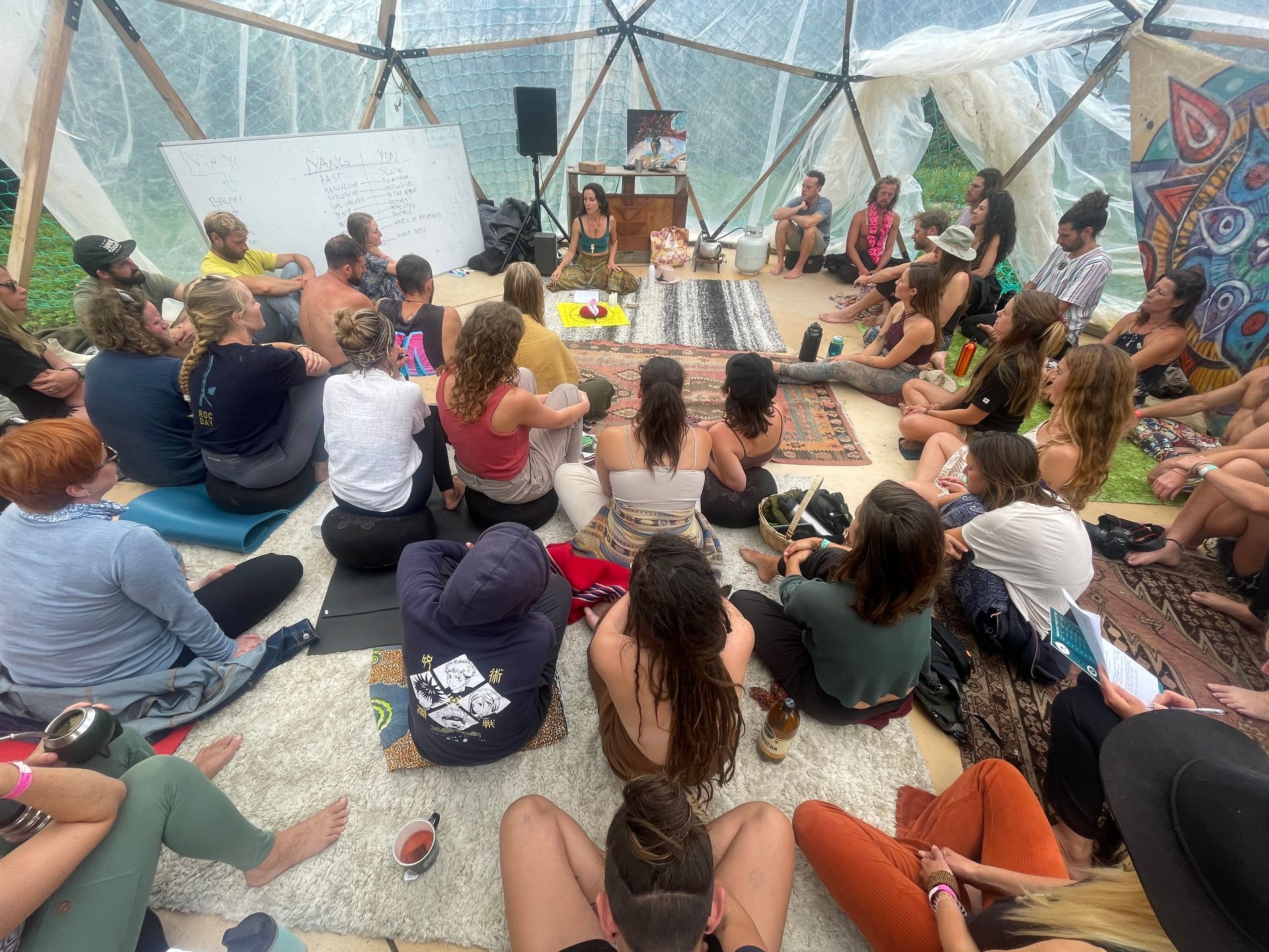 People sit in a geodesic dome, listening to a speaker, with a whiteboard and colorful art visible.