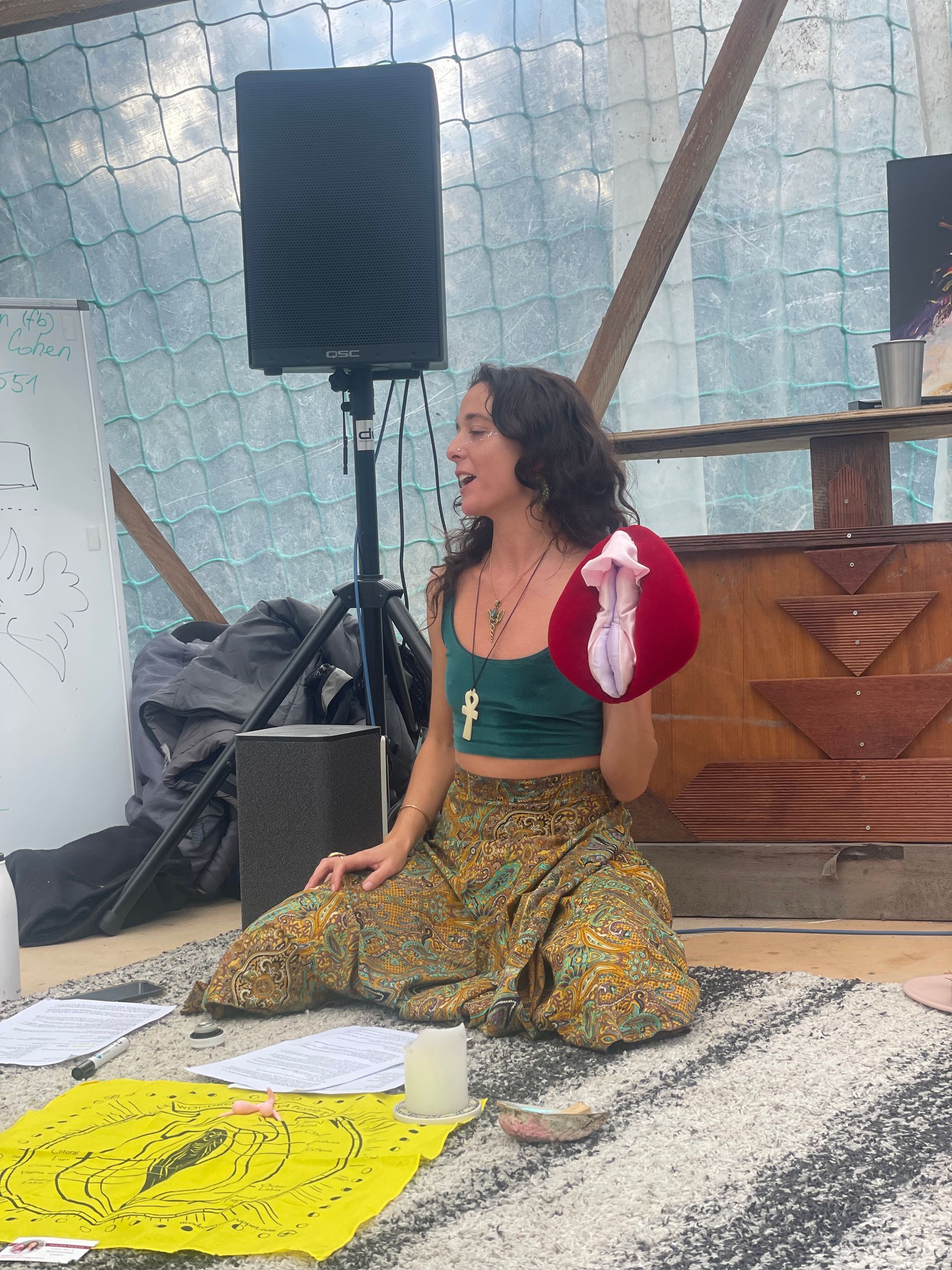 Woman seated holding a red vulva-shaped prop. She's giving a presentation with notes, speaker, in an outdoor setting.