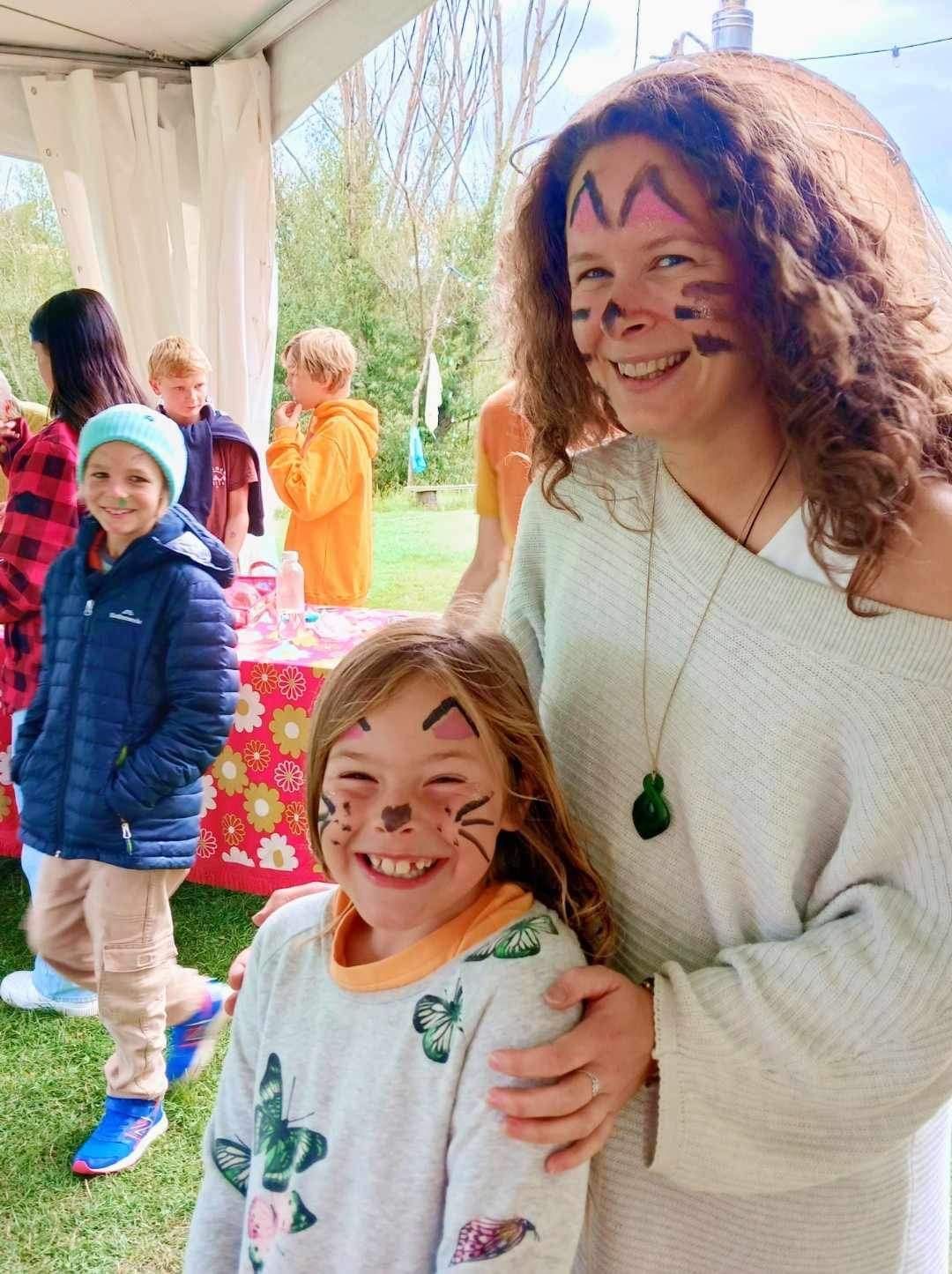 Woman and child with face paint smile at the camera, other people and a table are in the background.