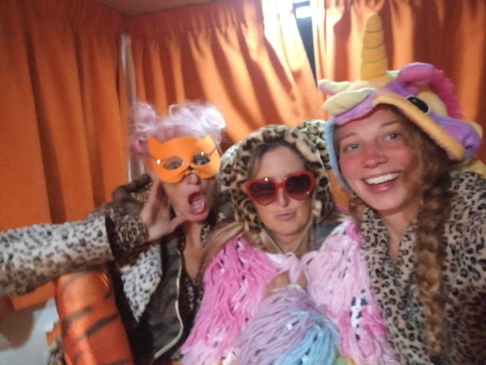 Three people in costumes, pink, leopard, and rainbow colors, making faces in front of orange curtains.