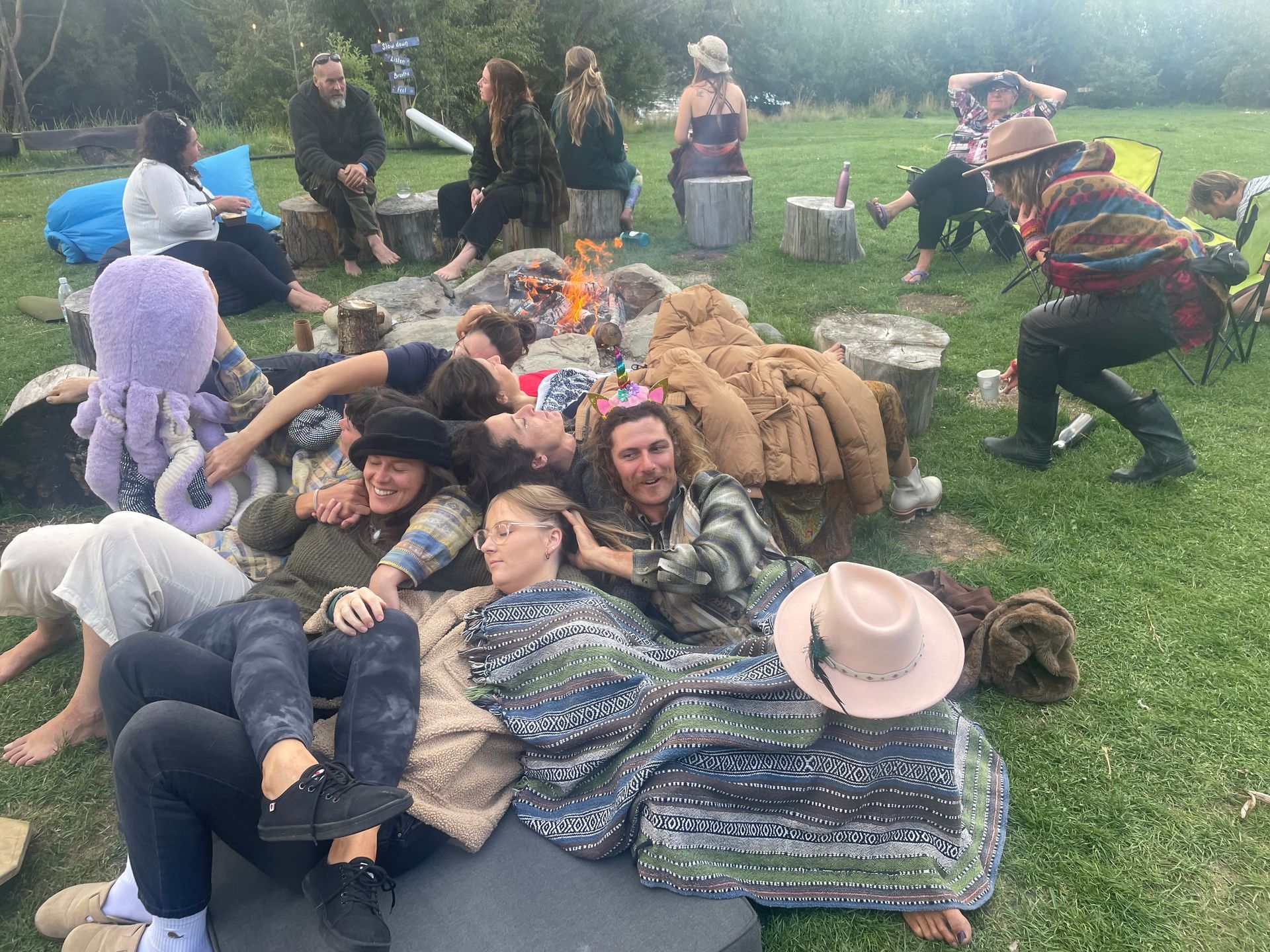 People gathered around a bonfire on grass. Some are lying down, others sitting, socializing in a casual outdoor setting.