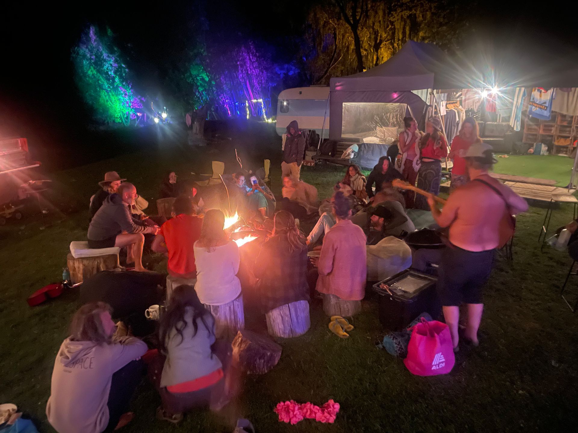 People gather around a bonfire at night.  Lights and a trailer visible in the background.