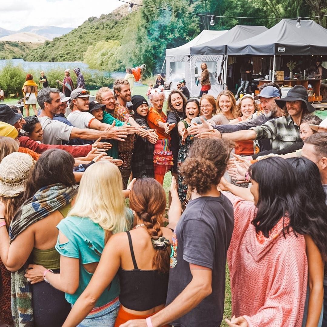 People in a circle with arms outstretched, smiles, outdoor setting with tents and lake.