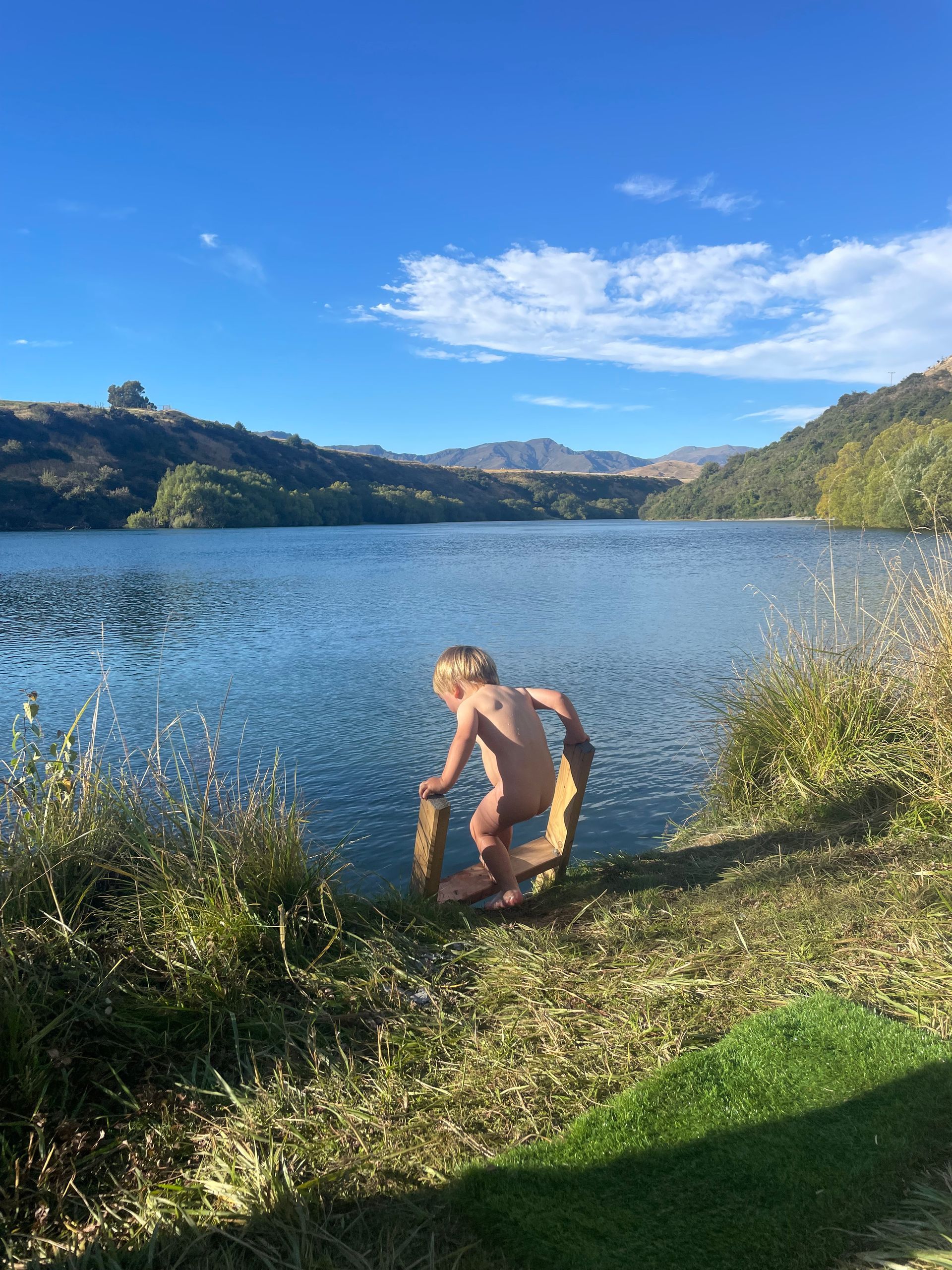 Child kneels at water's edge holding a coiled rope. A lake and hills are in the background under a blue sky.