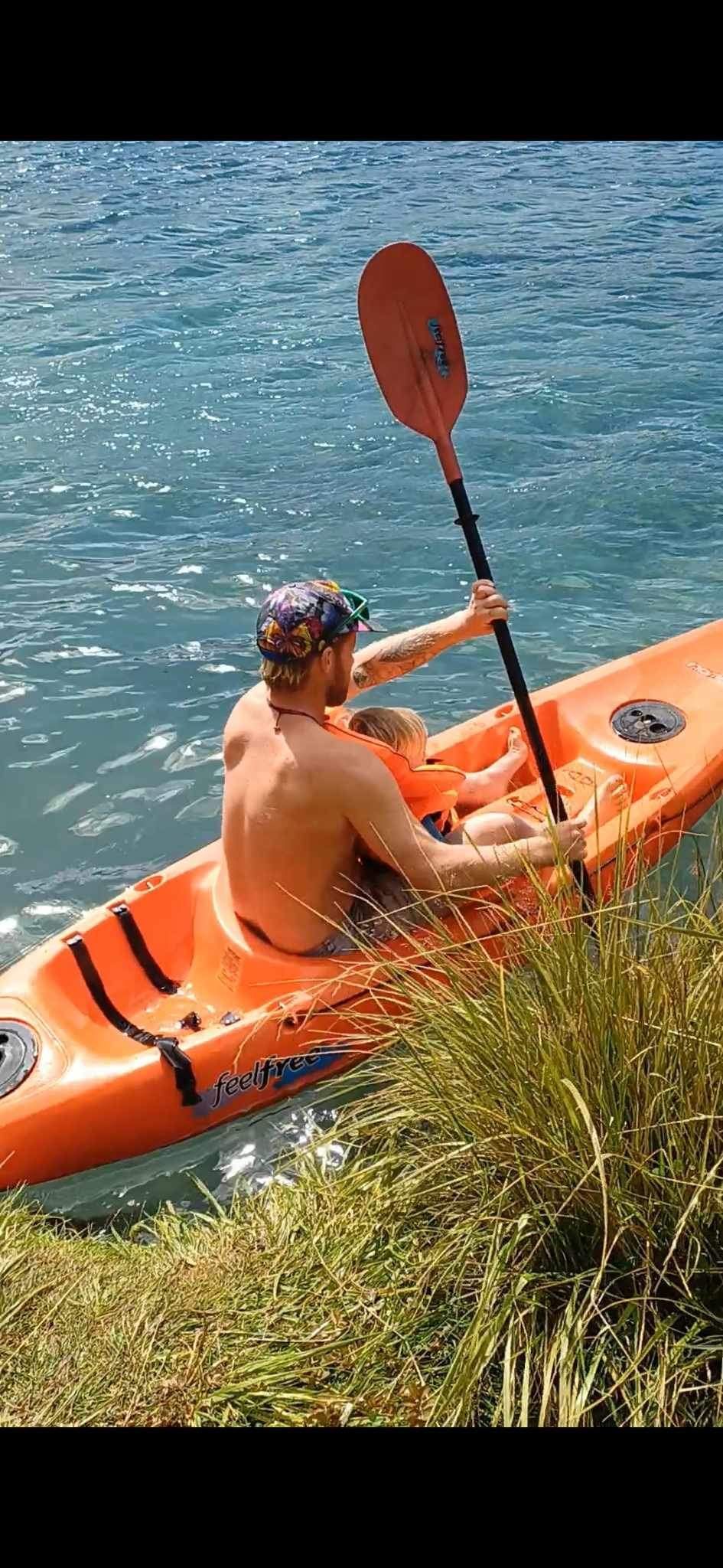 A person paddles an orange kayak on a lake with a child, near grassy shore.