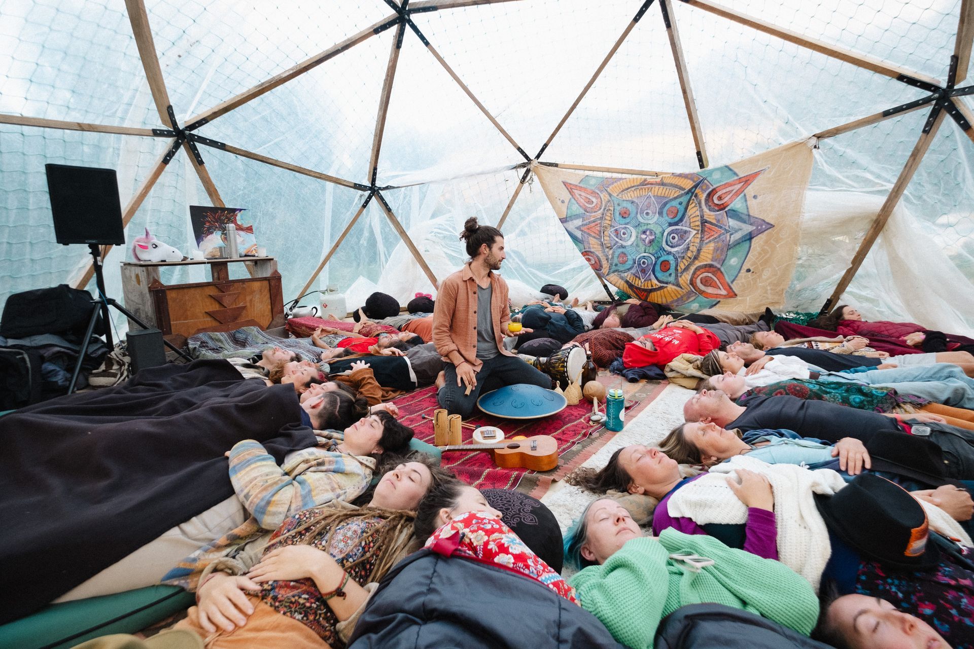 People lying on the floor in a dome-shaped structure, eyes closed, listening to a man with a drum.