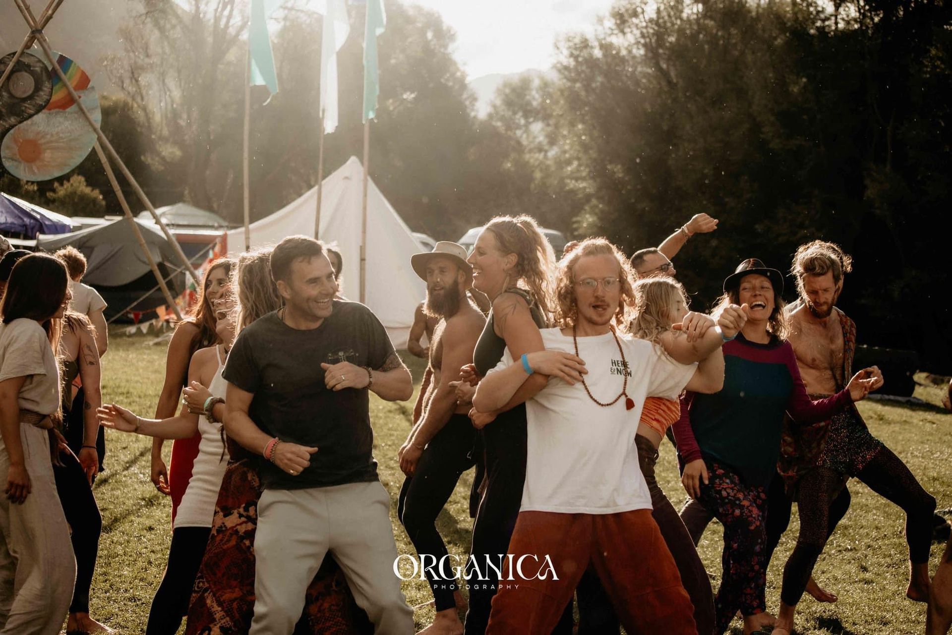 People dancing joyfully outdoors in a sunlit field with tents and flags in the background.