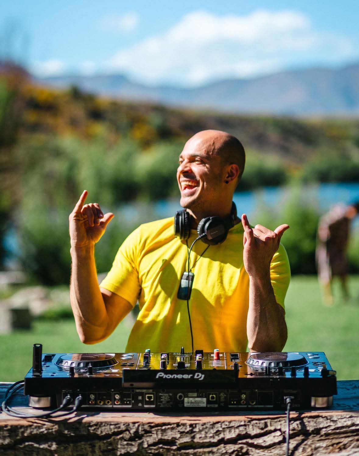 Man in yellow shirt, DJing outdoors, smiling, gesturing, with lake and trees in background.