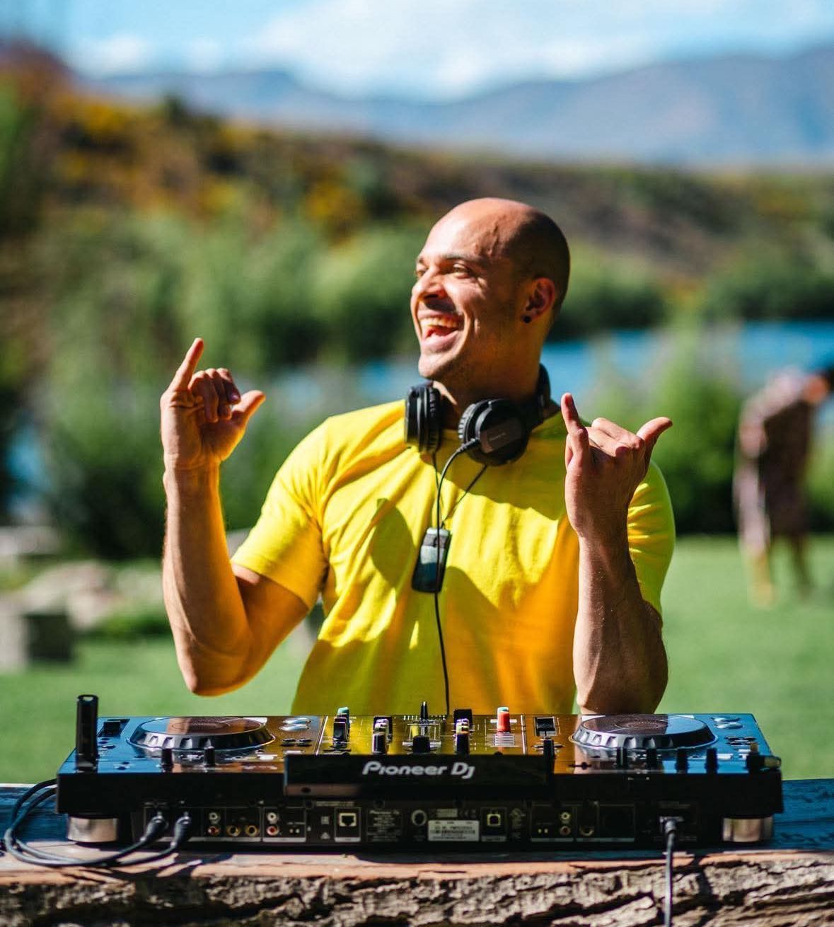 DJ in a yellow shirt with headphones smiles, giving hand gestures near his mixer, set outdoors near water.