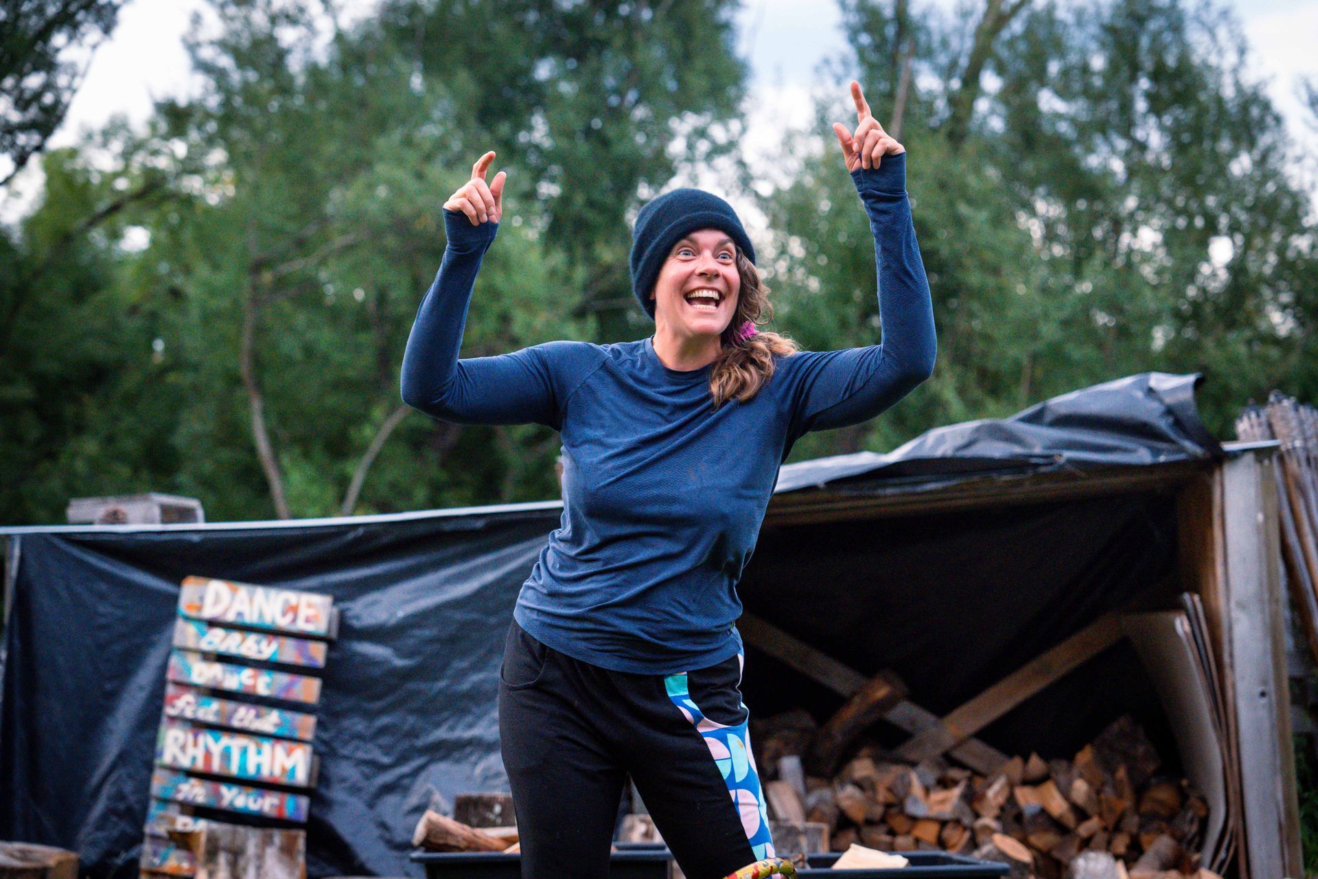 Woman with arms raised and fingers pointing up, smiling, wearing a hat, dark blue top, and black pants outdoors near a woodpile.