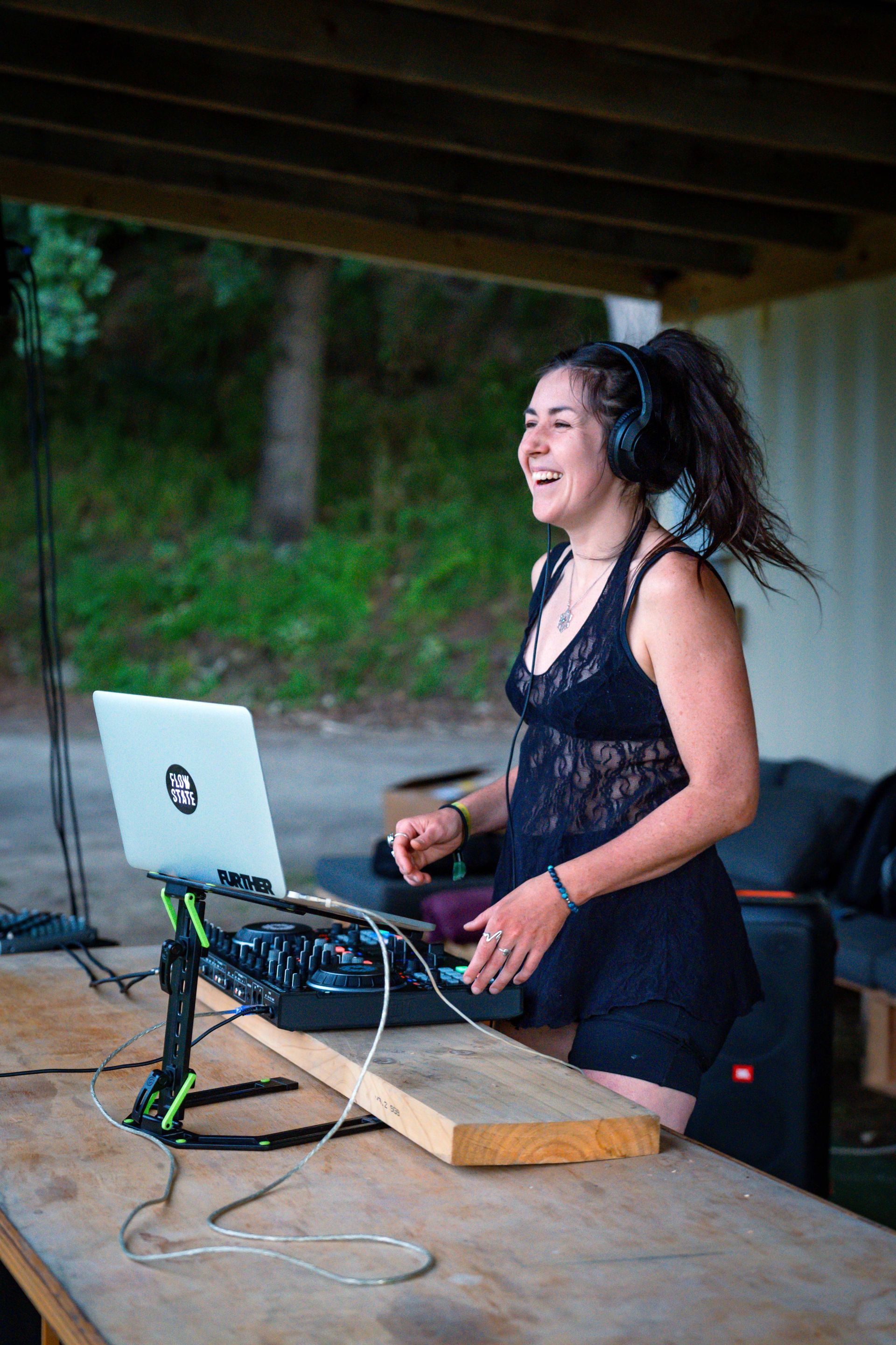 Woman DJing outdoors, laughing, wearing headphones and a black outfit, with laptop and equipment on a wooden table.