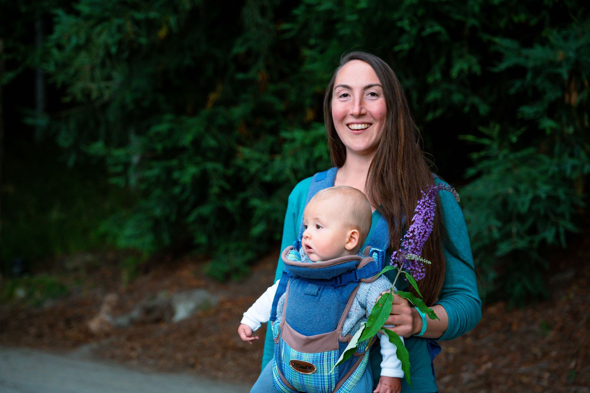 Woman holding baby in a carrier, smiling outside. Baby looks at the side. Green trees in the background.
