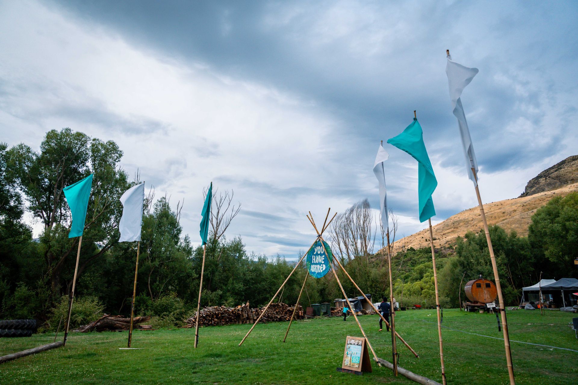 Green field with flags. Teal and white flags on wooden poles. Cloudy sky and trees in background.