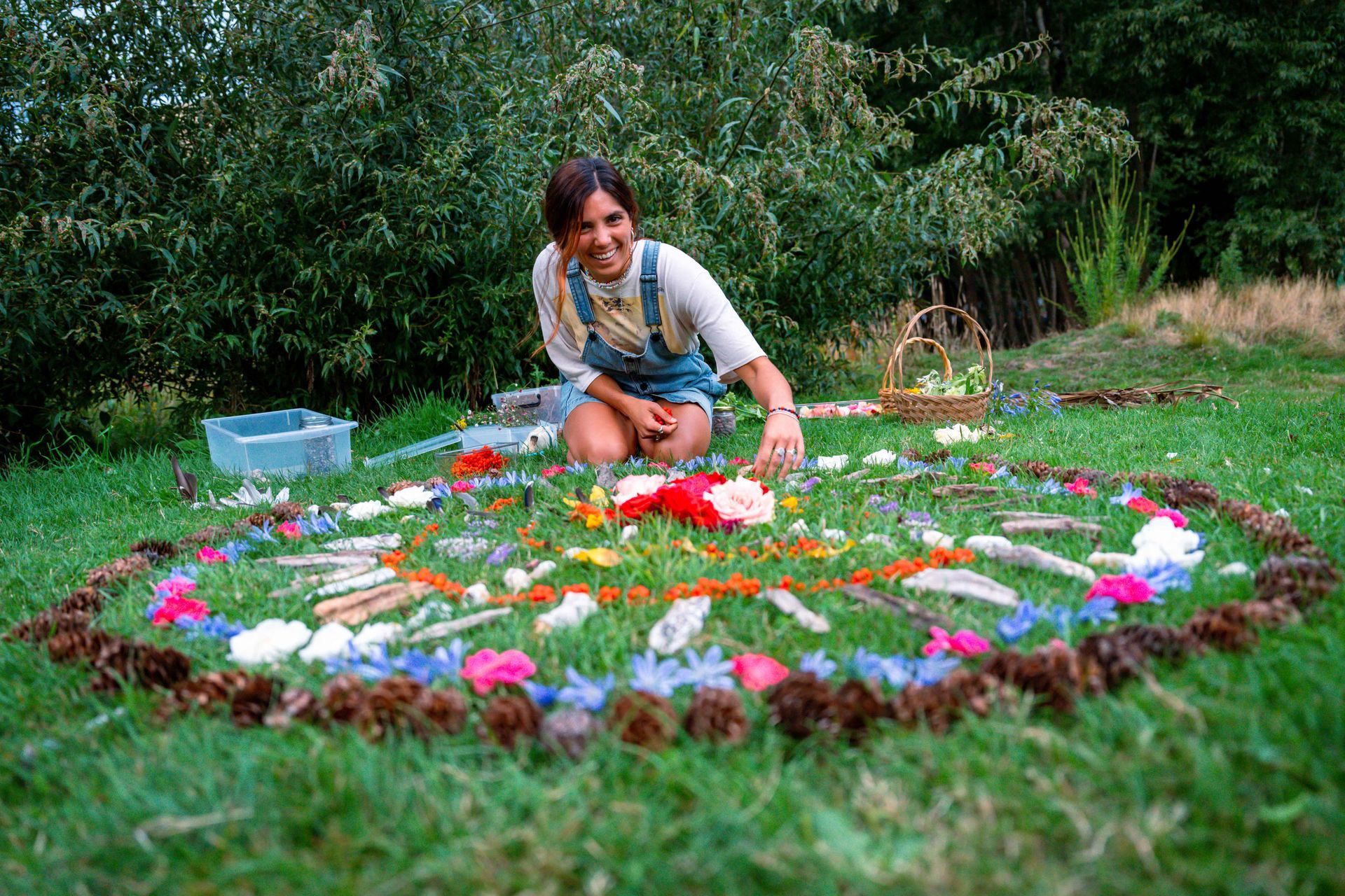 Woman creating a mandala on grass with flowers, pinecones, and feathers, smiling outdoors.