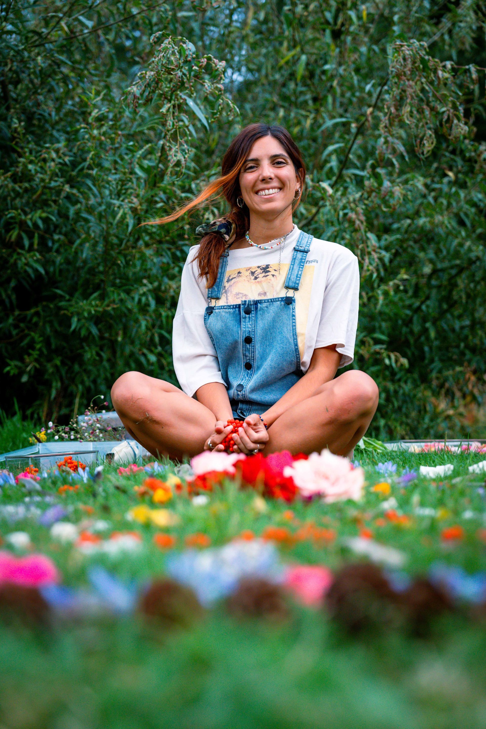 Woman seated cross-legged in a flower garden, smiling. Wearing overalls and white shirt. Outdoors with greenery in the background.